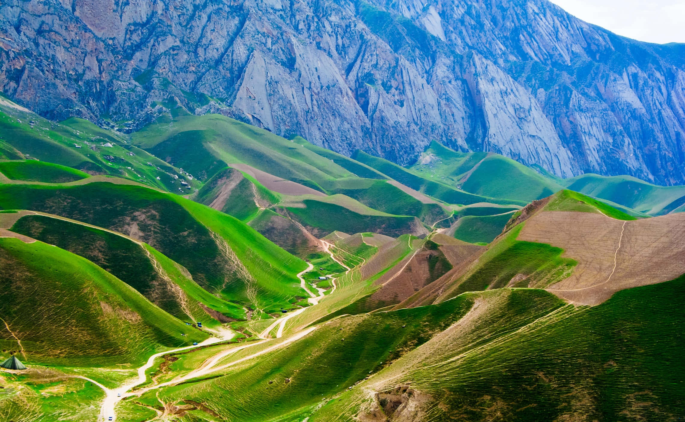 Afghanistan 桌布 壁紙 — 廣闊起伏的綠色山丘上蜿蜒的泥土路在明亮的日光下延伸至高聳崎嶇的山脈之下。
