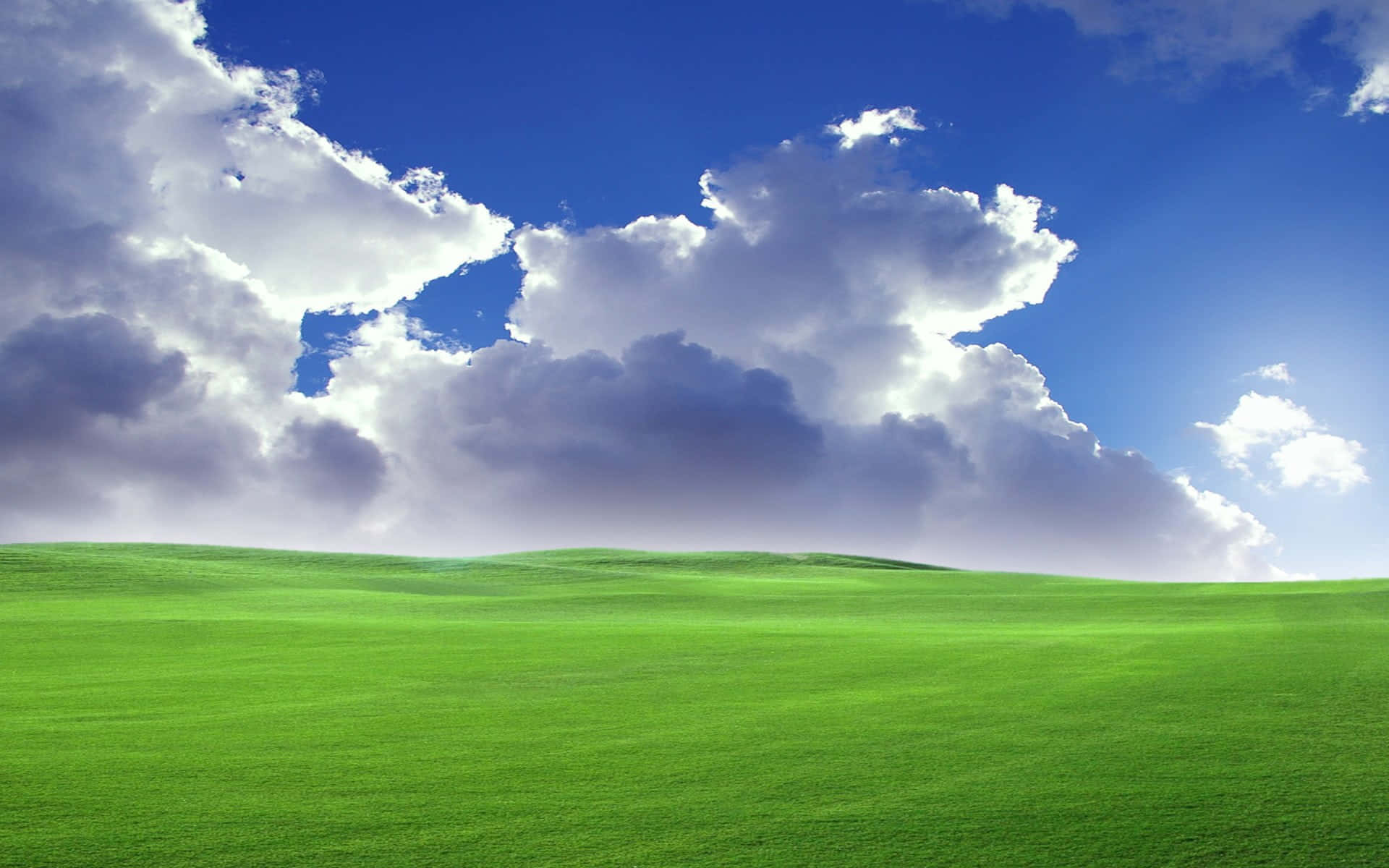 Green Grass 桌布 壁紙 — 廣闊起伏的綠色山丘在戲劇性的天空下延伸，天空充滿了蓬鬆的白色和灰色雲朵，背景是深藍色。