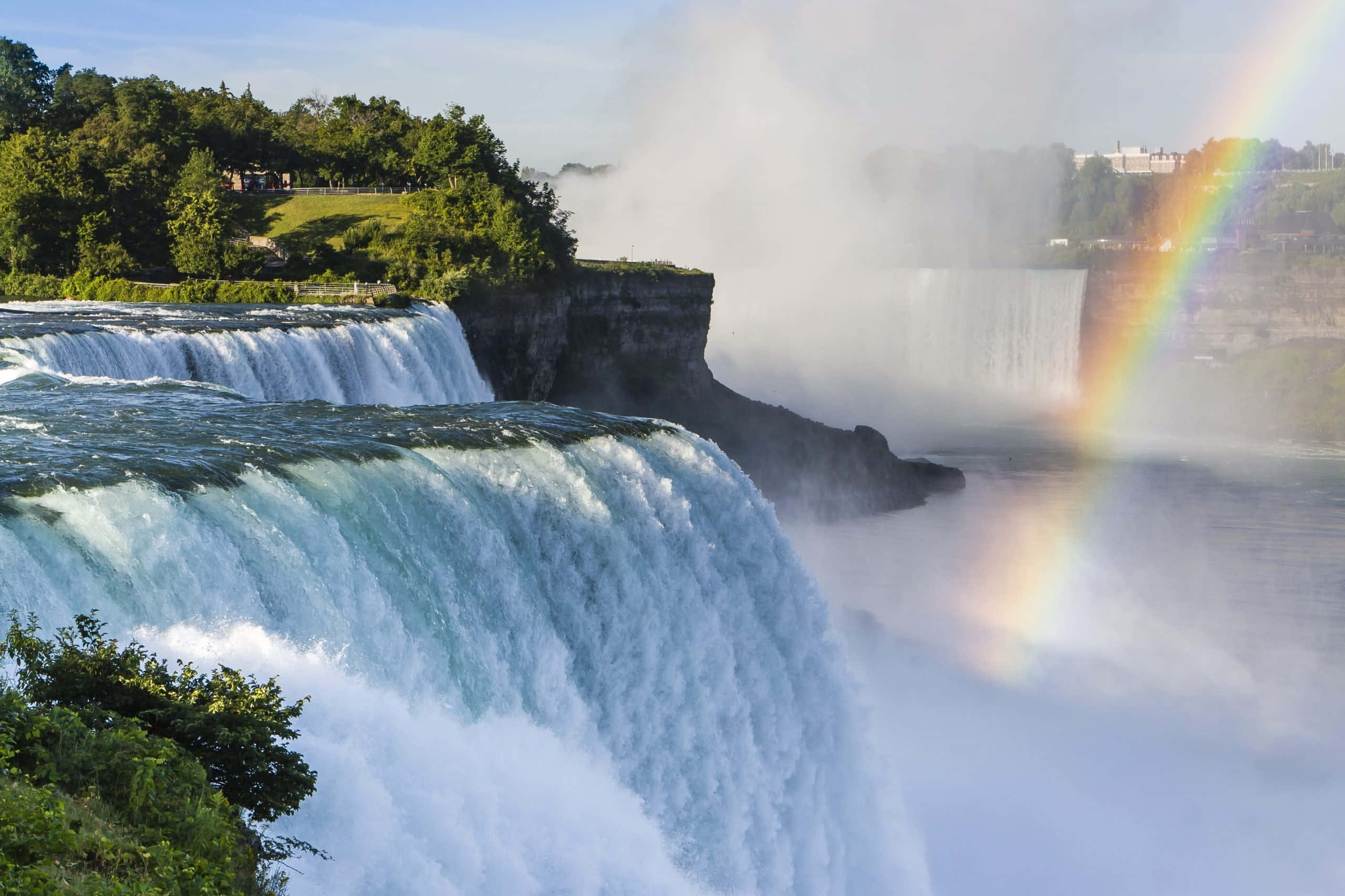 Niagara Falls 桌布 壁紙 — 強勁的層疊瀑布產生水霧，在晴朗的藍天和茂密的綠植中形成鮮豔的彩虹。