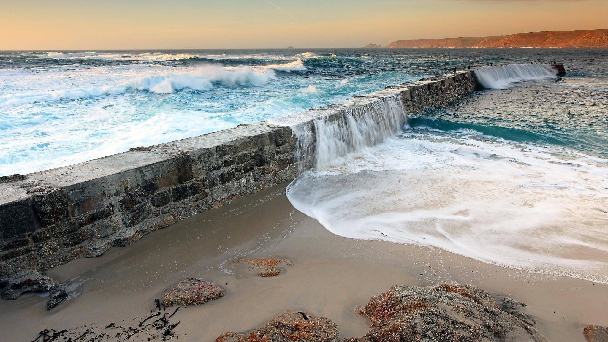 海洋 桌布 壁紙 — 強勁的海浪在金色日落時分衝擊著堅固的石頭防波堤，將溫暖的光芒灑在泡沫翻騰的浪花和沙灘上。
