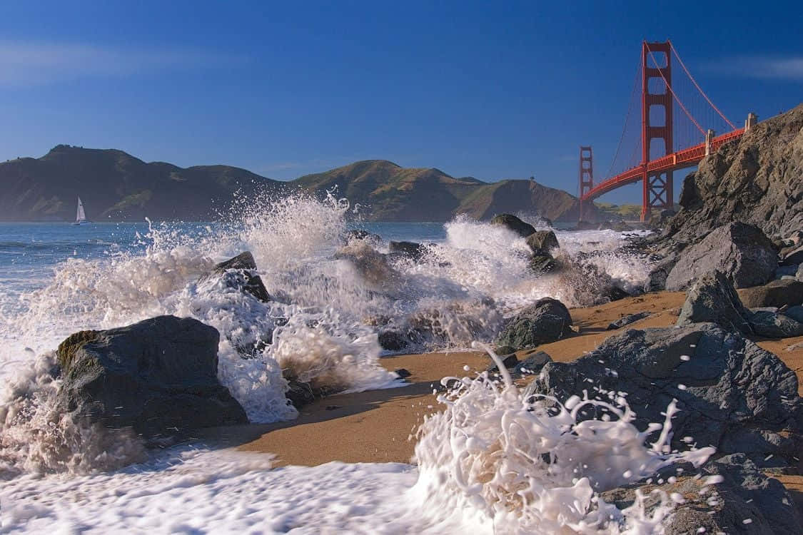 waves 桌布 壁紙 — 強勁的海浪拍打崎嶇的岩石，標誌性的金門大橋在清澈的藍天下聳立在背景中。