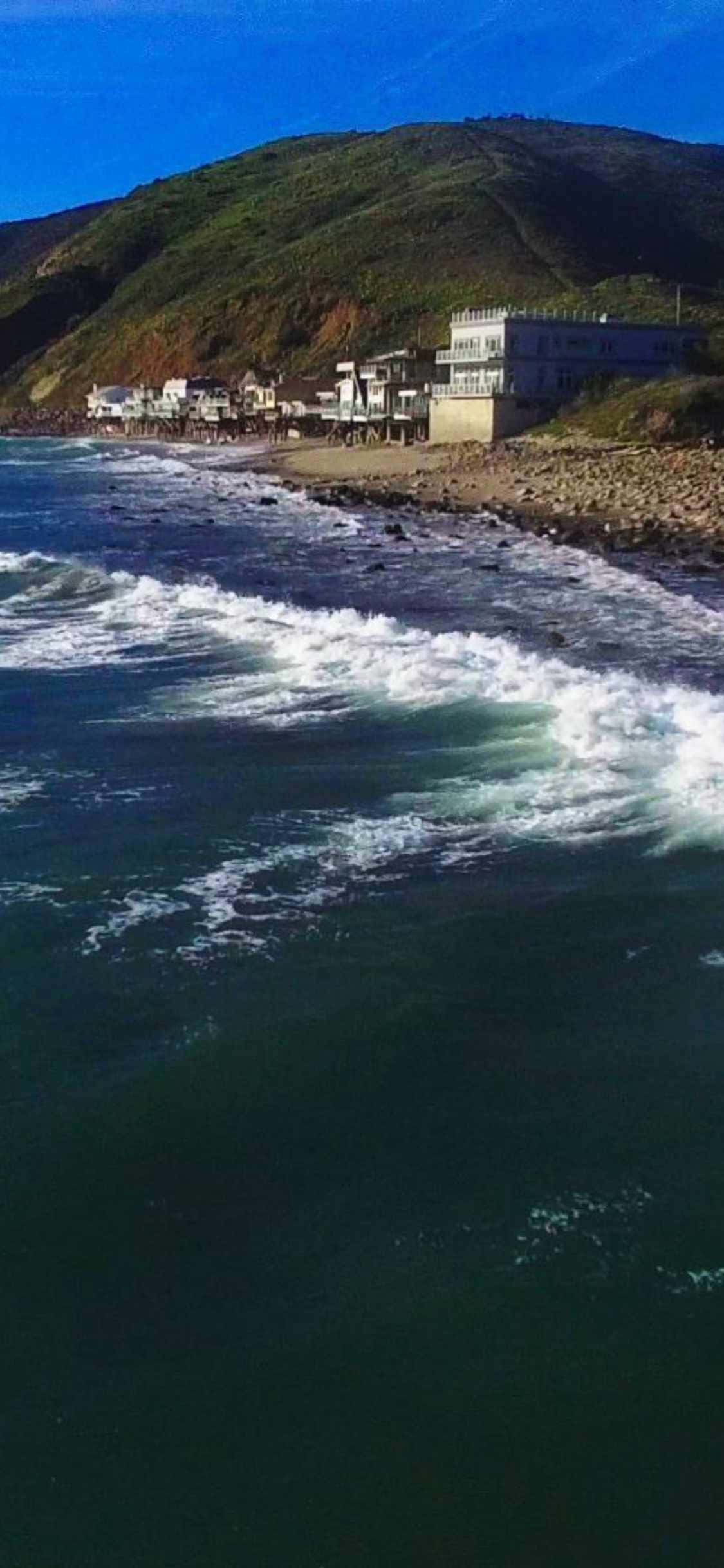 iPhone Xs Malibu 桌布 壁紙 — 強勁的海浪拍打著岩石海岸，下方是白色建築物依附在綠色的山坡上，上方是清澈的藍天。