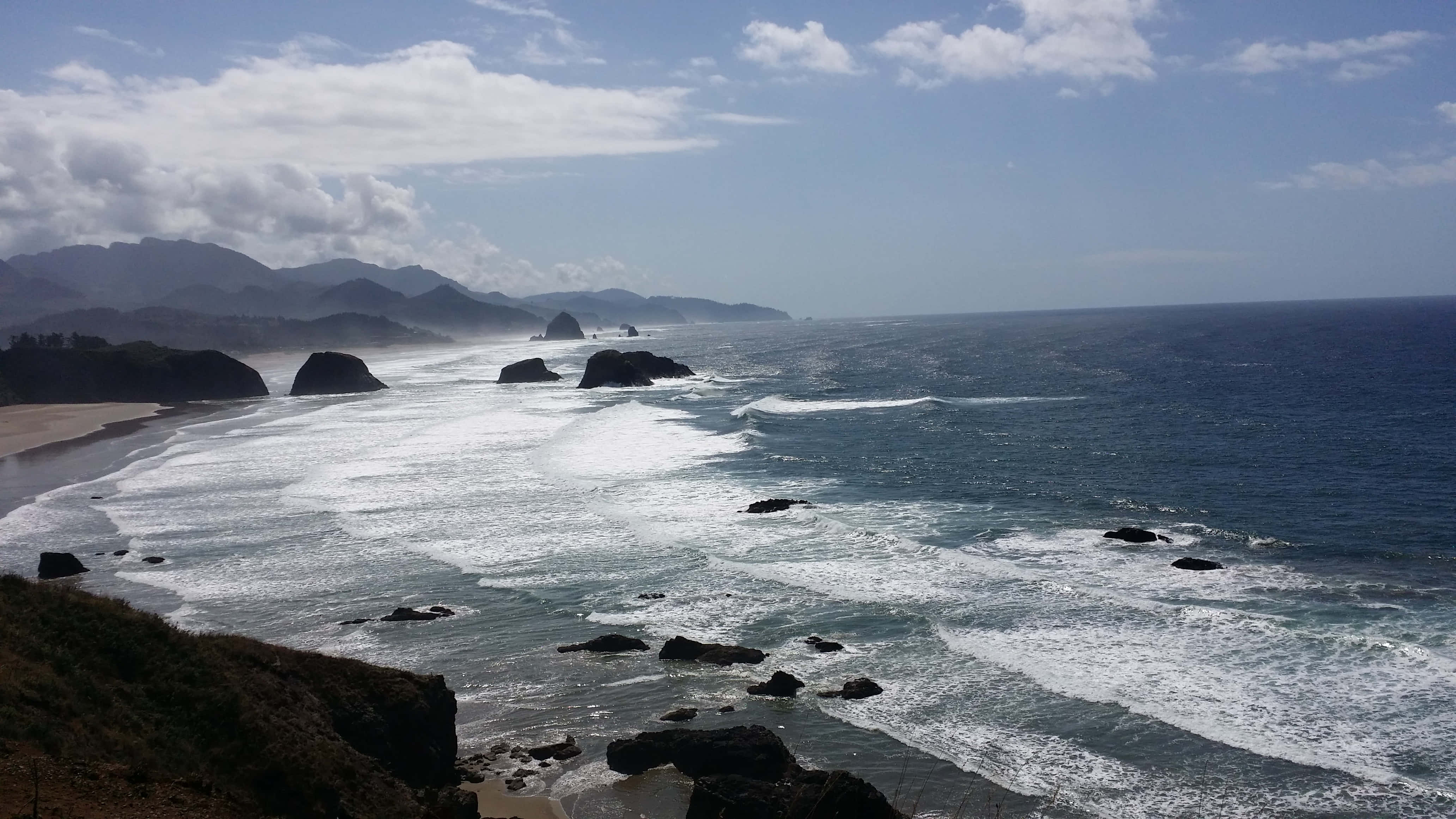 Oregon Coast 桌布 壁紙 — 強勁的海浪拍打著崎嶇的黑色岩石，沿著蜿蜒的海岸線，天空部分多雲。