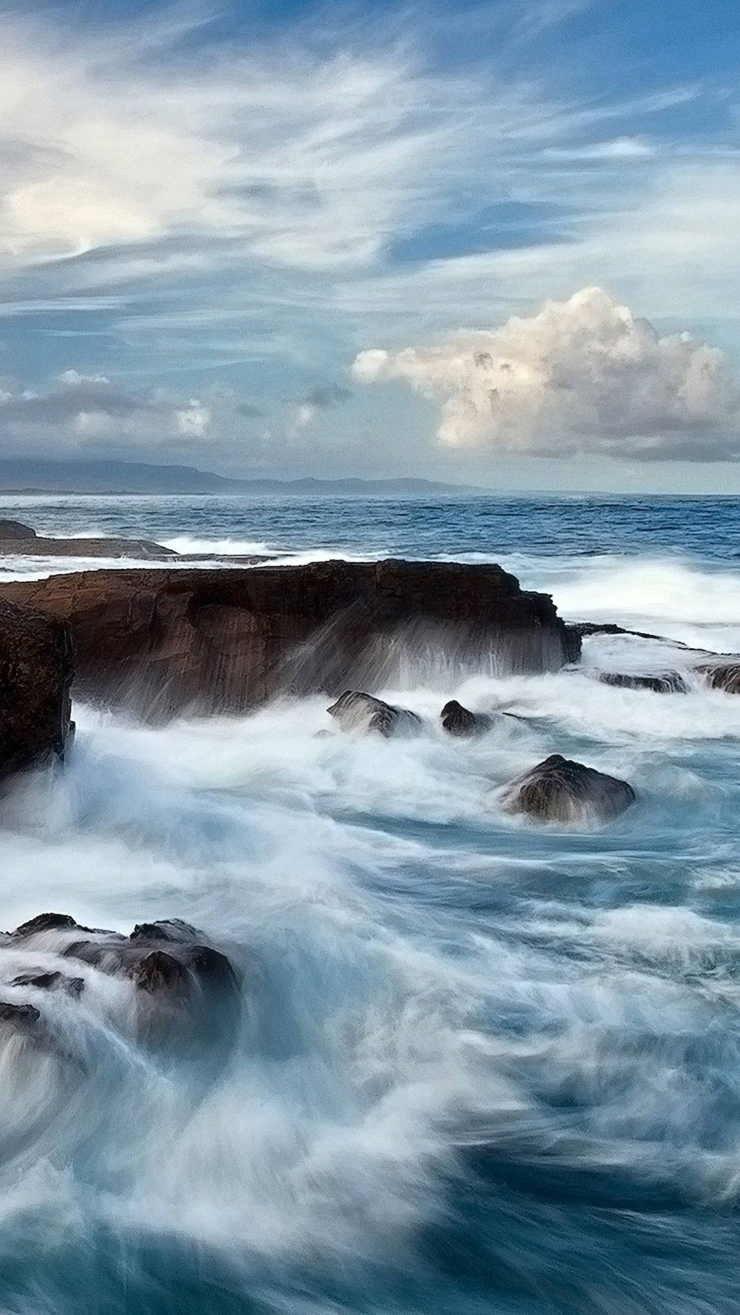 Ocean iPhone 桌布 壁紙 — 強勁的海浪拍打著深色的火山岩，廣闊的天空中飄著縷縷雲彩，遠山隱約可見。