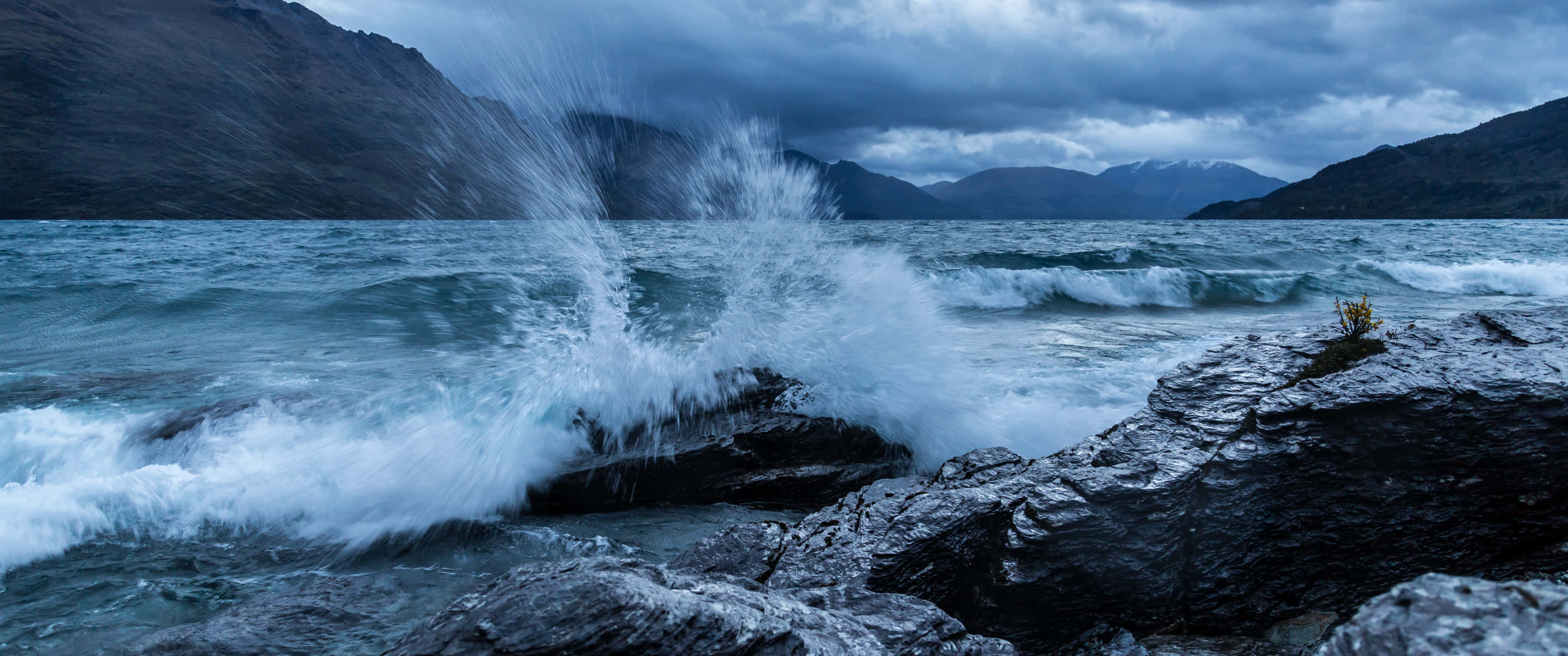 waves 桌布 壁紙 — 強勁的海浪拍打著黑暗的鋸齒狀岩石，天空灰暗風暴，遠山隱約可見。