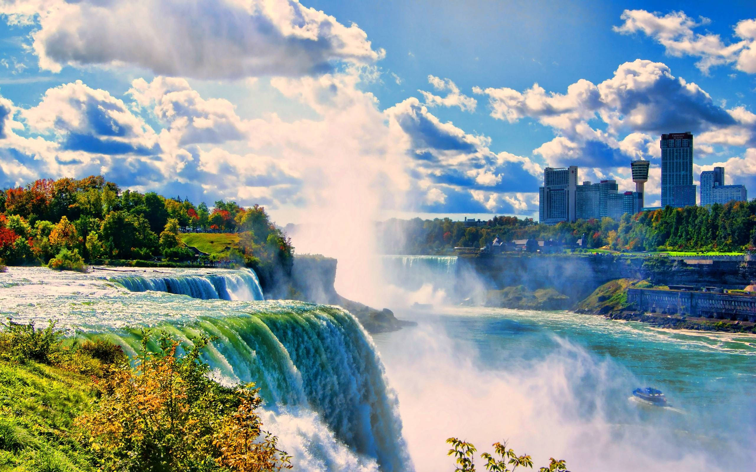 niagara 桌布 壁紙 — 強勁的藍綠色水流從尼亞加拉瀑布傾瀉而下，在明亮的藍天、蓬鬆的白雲和背景的城市天際線下形成水霧。
