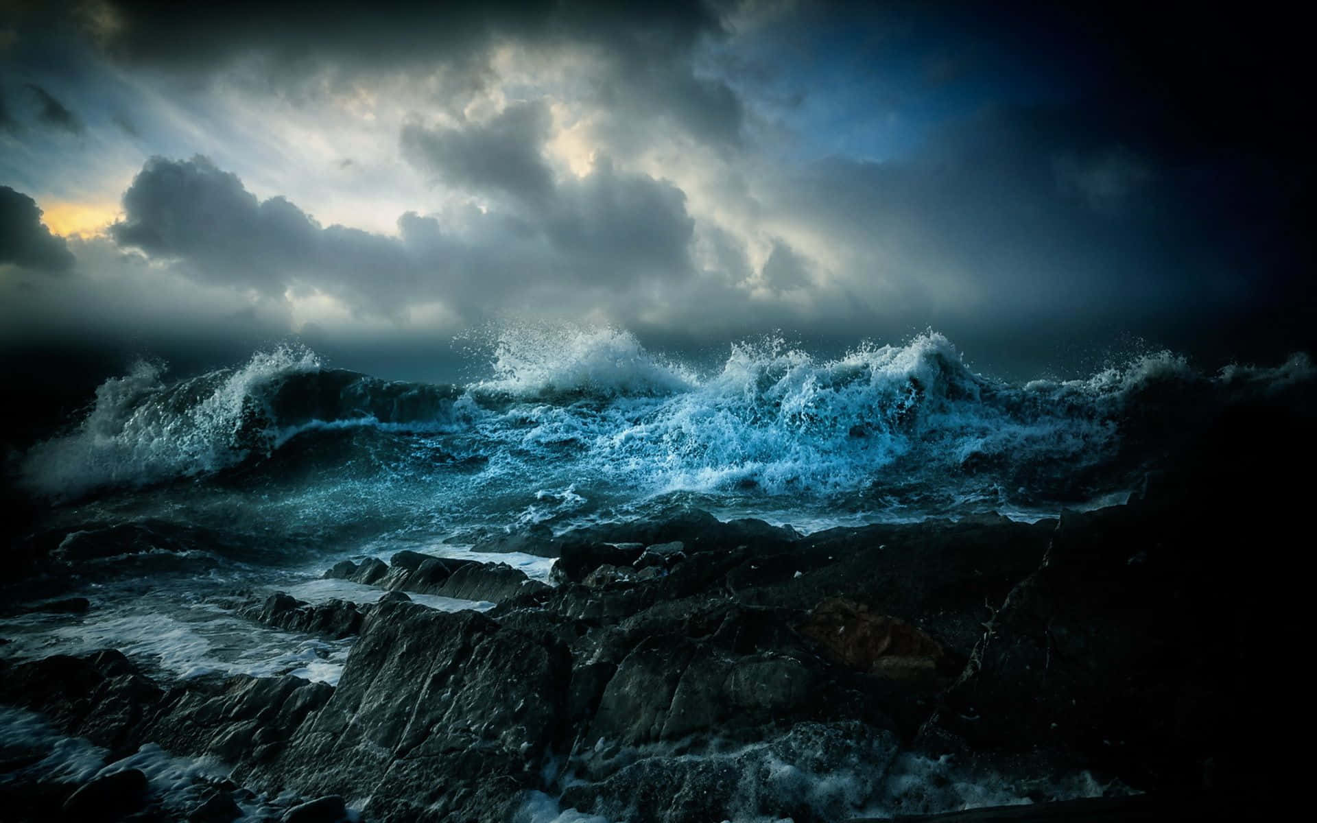 Ocean Storm 桌布 壁紙 — 強勁的藍色波浪在暴風雨的天空下，以戲劇性的背光衝擊著黑暗的岩石。