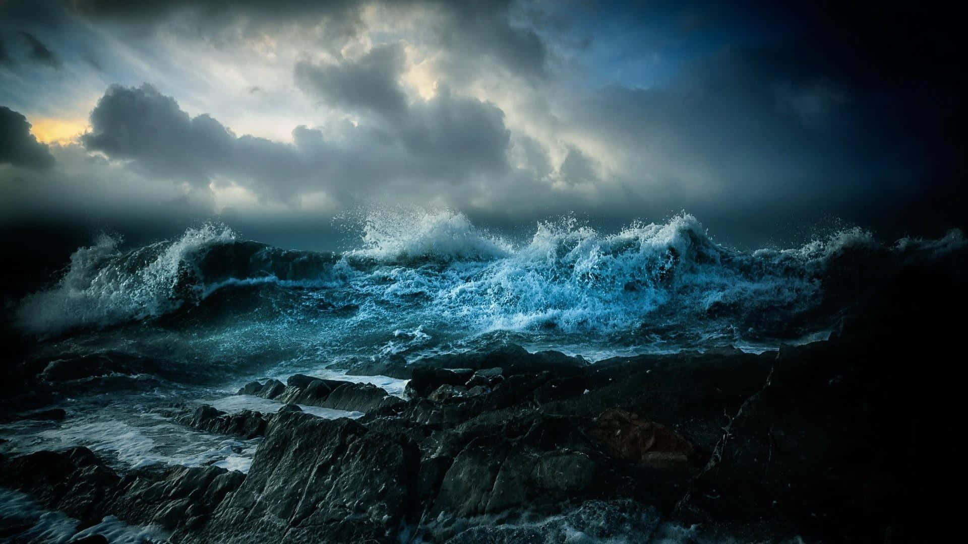 Stormy 桌布 壁紙 — 強勁的藍色波浪在暴風雨的天空下撞擊黑暗的岩石，伴隨著戲劇性的光線。