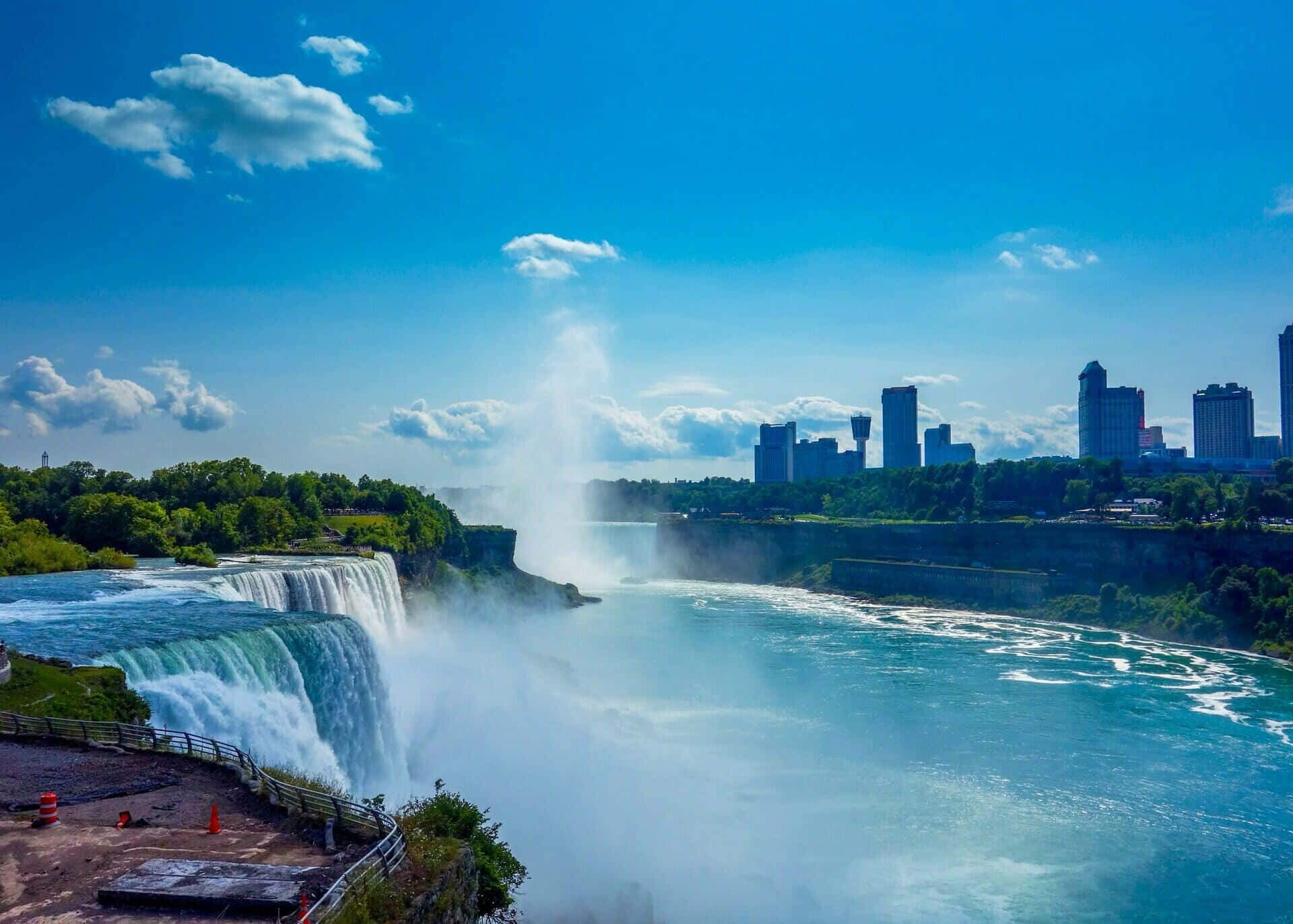 Niagara Falls 桌布 壁紙 — 強大的尼亞加拉瀑布在明亮的藍天、散落的雲朵和遠處的城市天際線下奔流而下。