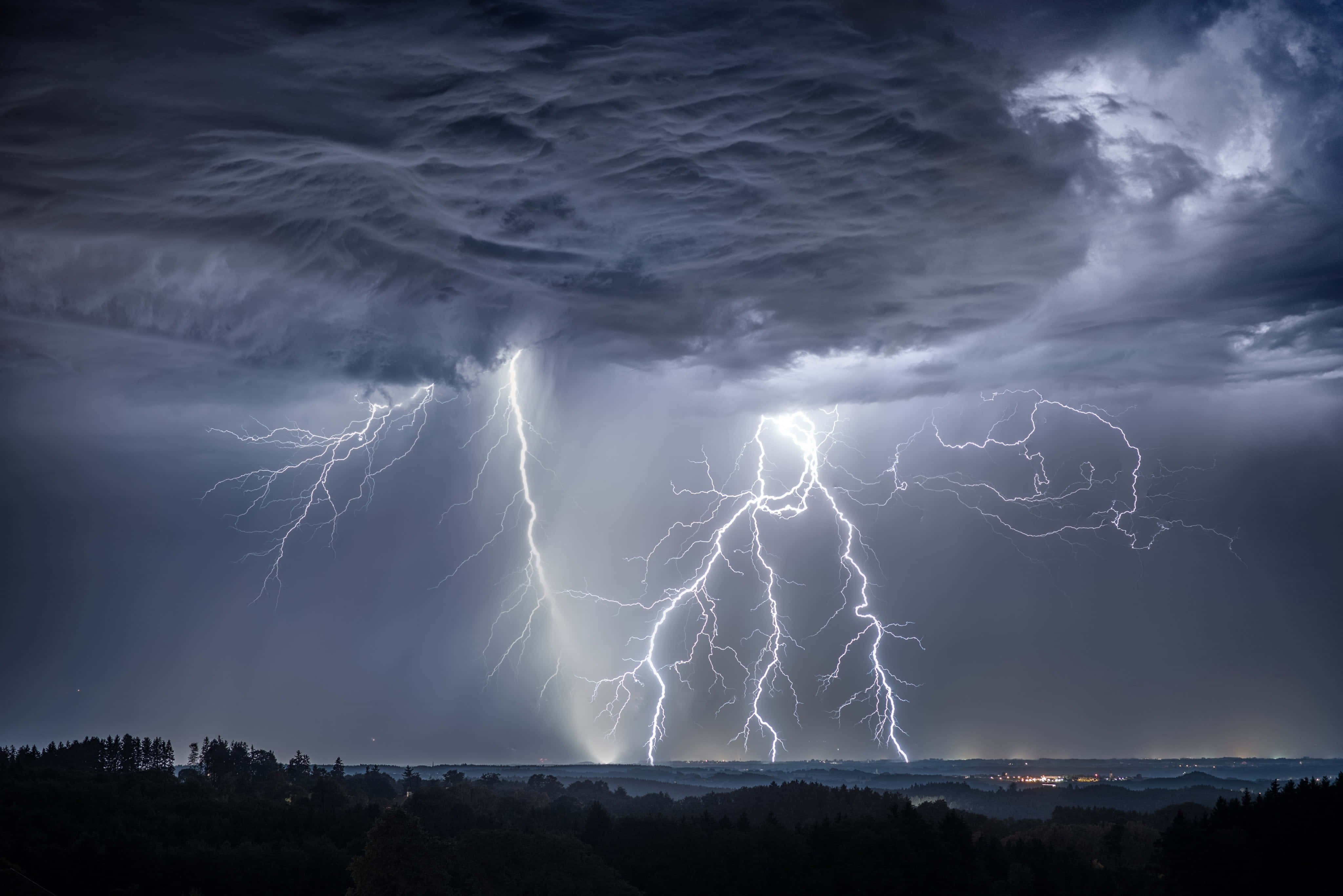 暴風 桌布 壁紙 — 強大的閃電劃破暴風雨的夜空，下方是黑暗的森林地平線和遠處的城市燈光。