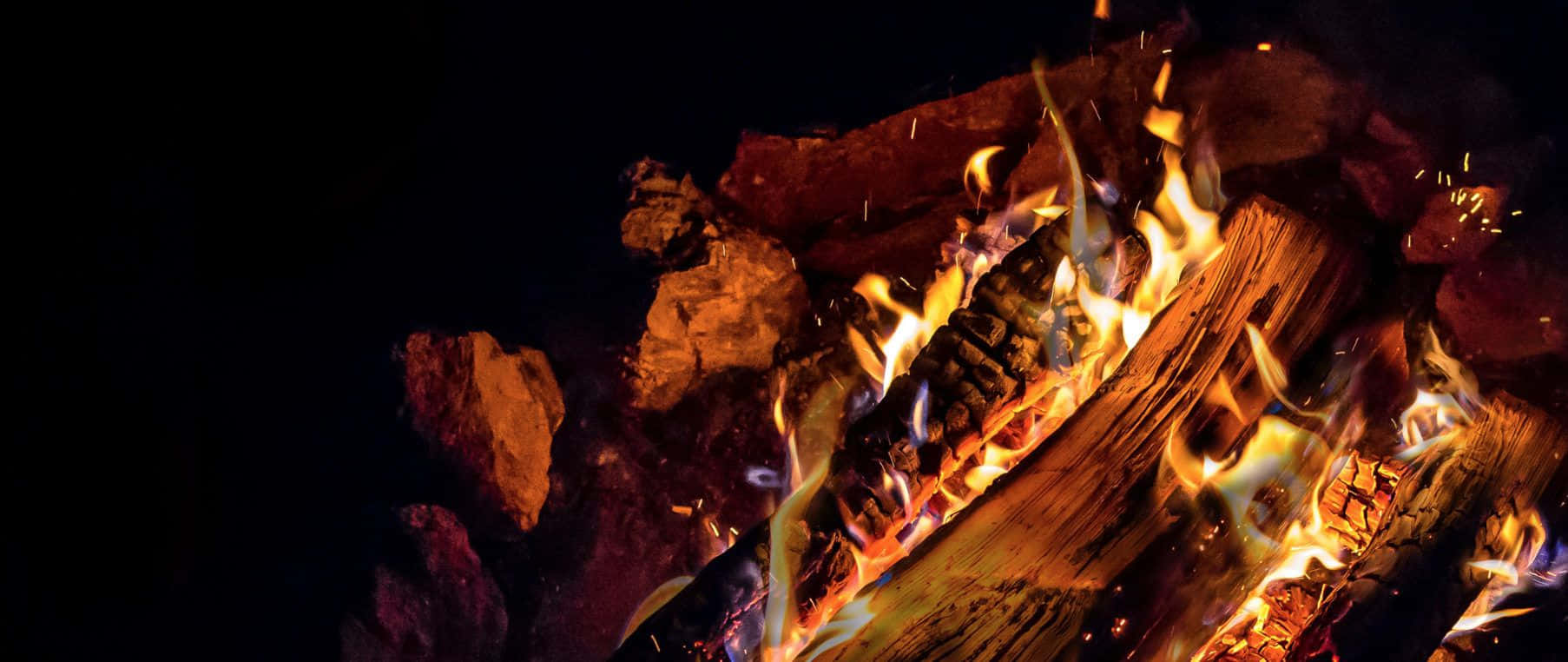 Campfire 桌布 壁紙 — 強烈的火焰從燃燒的木頭中躍出，周圍是黑色的岩石，背景是漆黑的夜空。