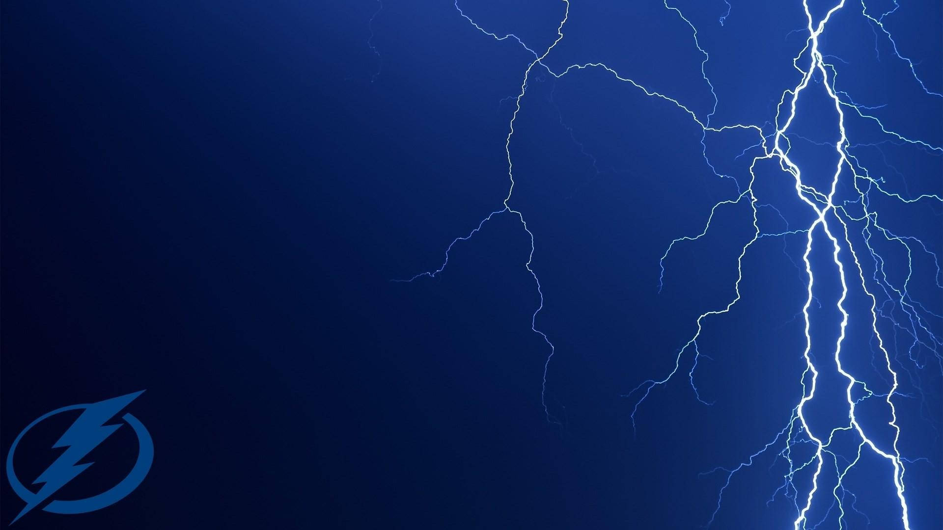 坦帕灣閃電 桌布 壁紙 — 強烈的白色閃電在深藍色暴風雨天空中劈啪作響，角落有一個微妙的閃電標誌。