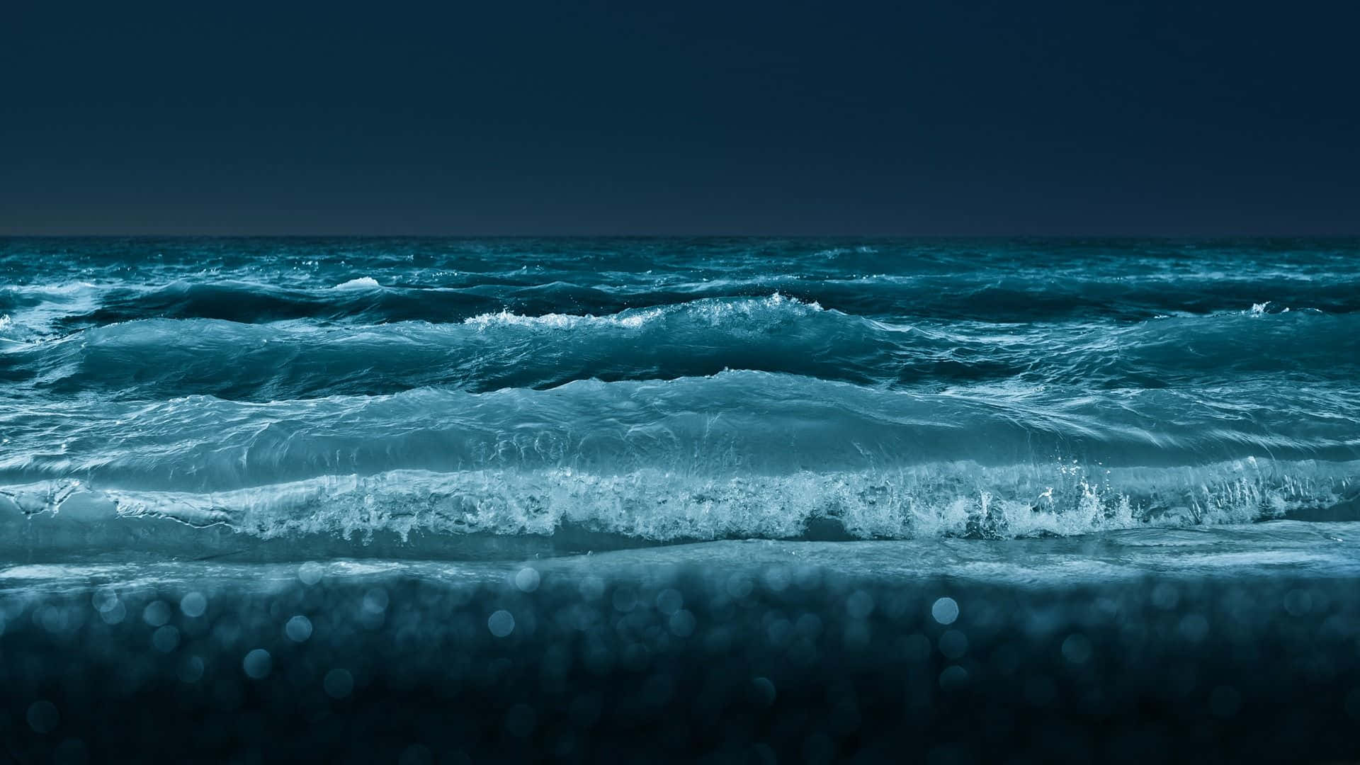 Ocean Waves 桌布 壁紙 — 強烈的藍綠色波浪在陰沉的夜空下拍打著黑暗的海岸，前景帶有散景效果。