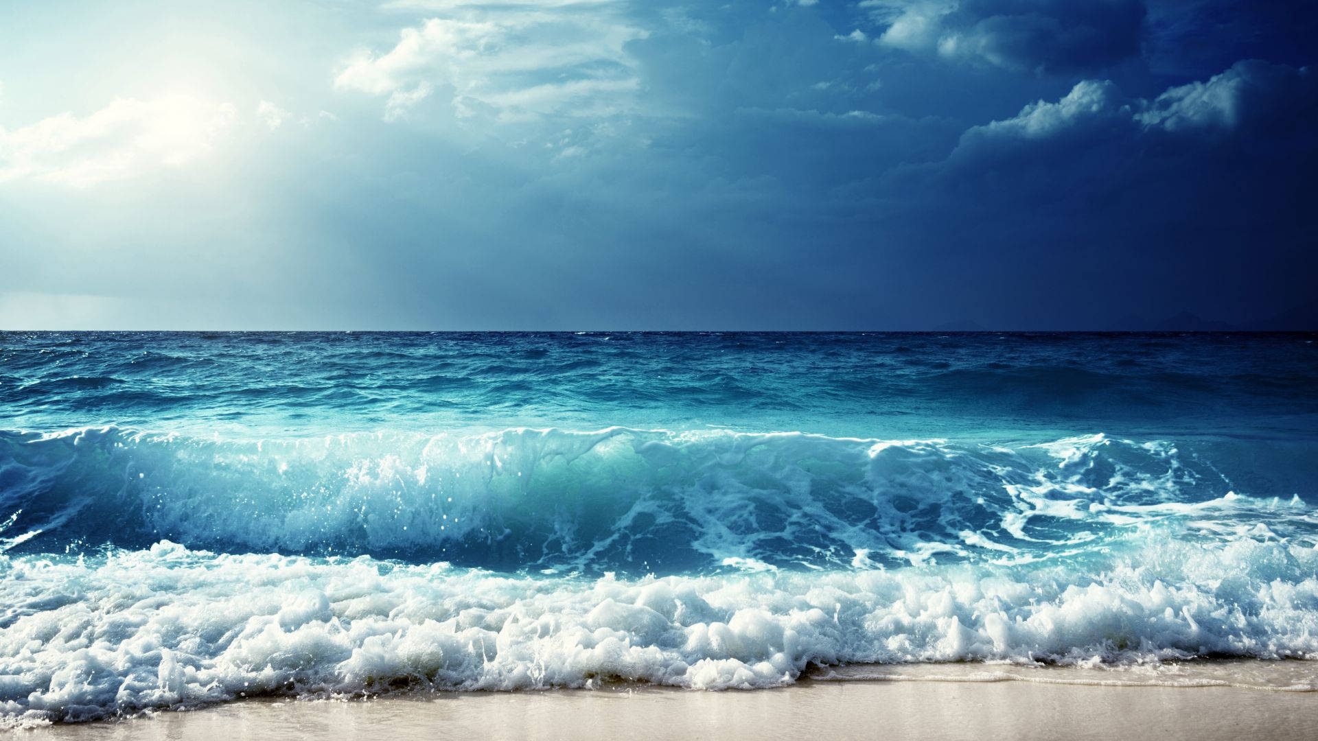 waves 桌布 壁紙 — 強烈的藍綠色海浪拍打著沙灘，天空烏雲密佈，陽光穿透雲層，場面壯觀。