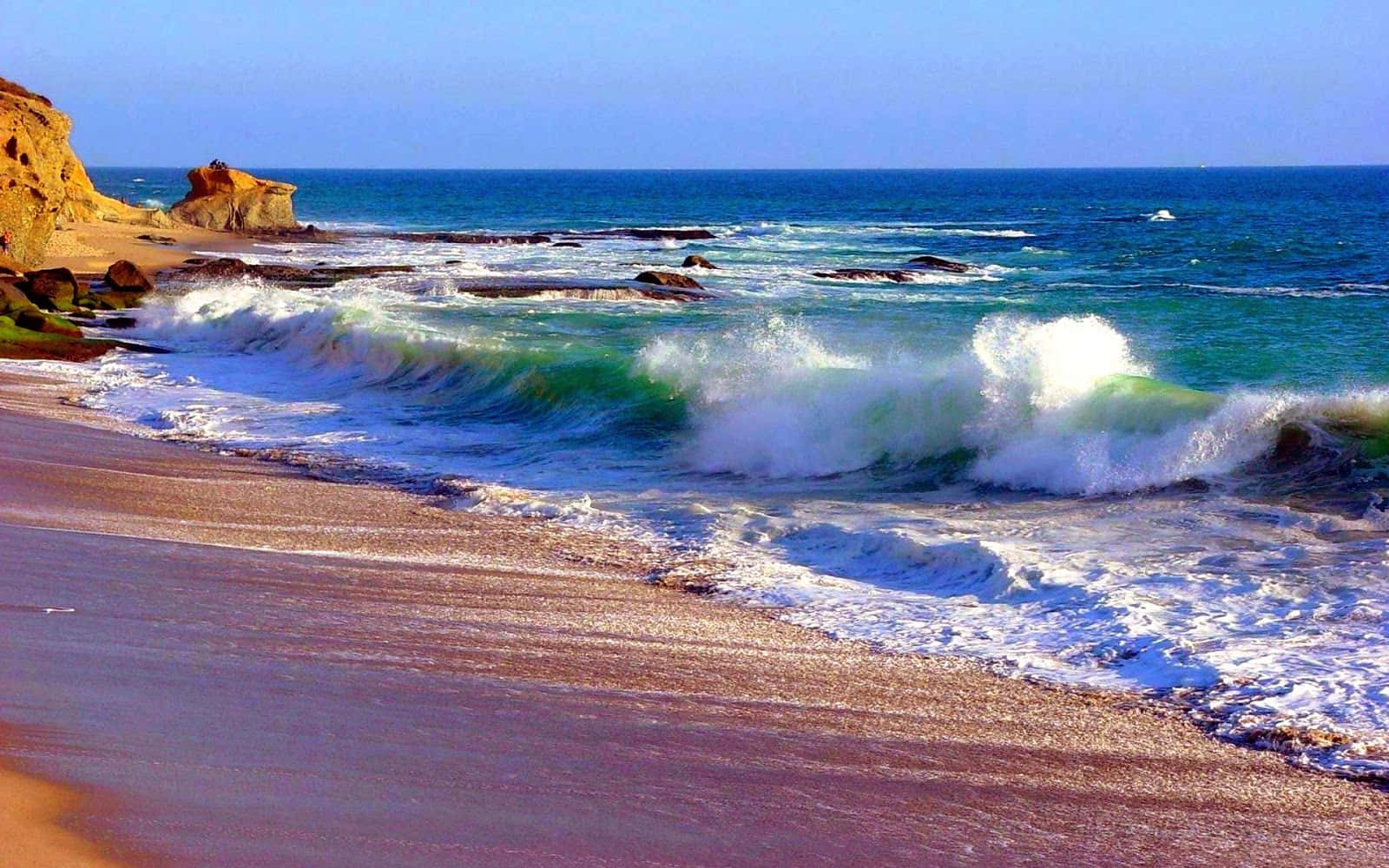 Ocean Waves 桌布 壁紙 — 強烈的藍綠色海浪拍打著陽光明媚的沙灘，旁邊是崎嶇的金色懸崖，上方是清澈的藍天。