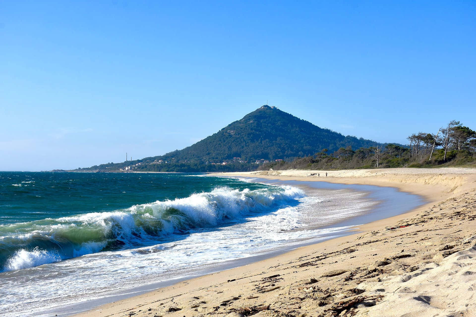 Portuguese Beach 桌布 壁紙 — 強烈的藍綠色海浪拍打陽光明媚的沙灘，背景是樹木繁茂的山丘，天空清澈湛藍。
