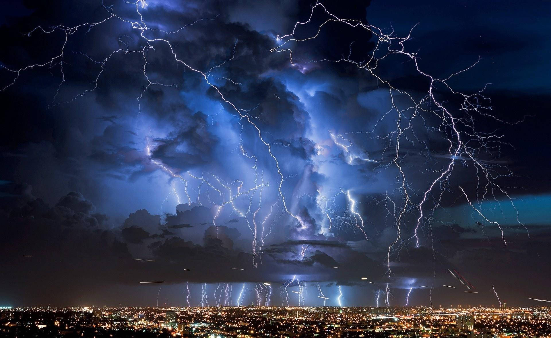 閃電 桌布 壁紙 — 強烈的閃電在暴風雨的夜空中劈啪作響，下方是燈火輝煌的廣闊城市景觀。