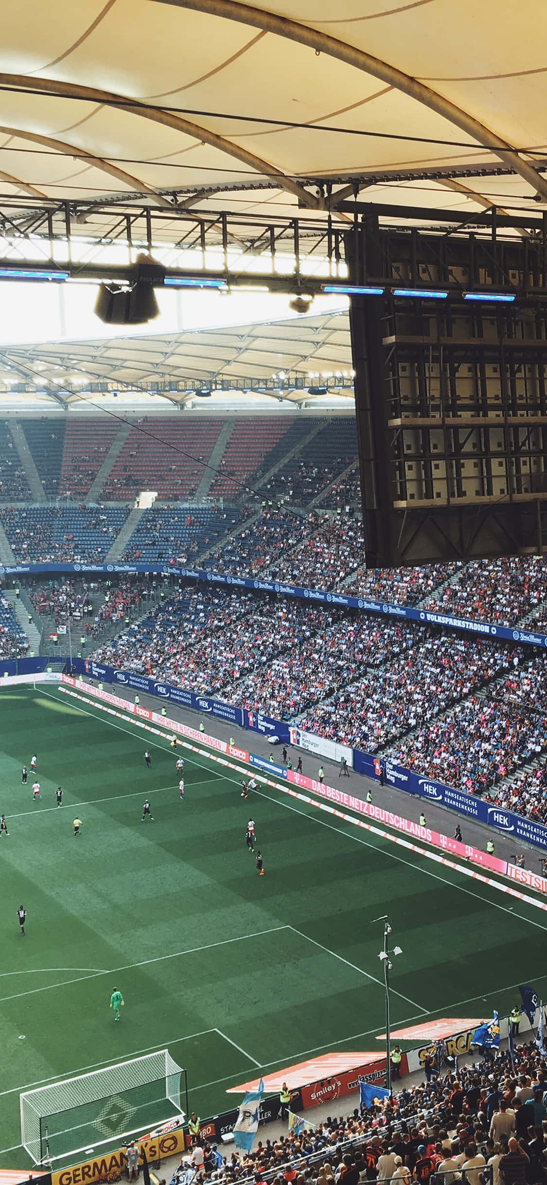 Soccer Stadium 桌布 壁紙 — 彎曲屋頂結構下，球員在充滿活力的綠色草坪上的擁擠足球場的鳥瞰圖。