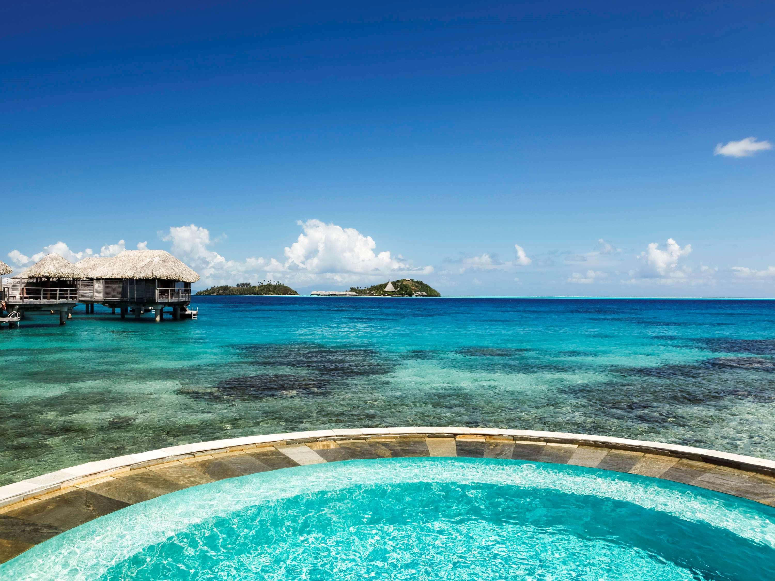 Bora Bora Beach 桌布 壁紙 — 彎曲的無邊際泳池俯瞰著碧綠的海水和水上平房，天空清澈湛藍。