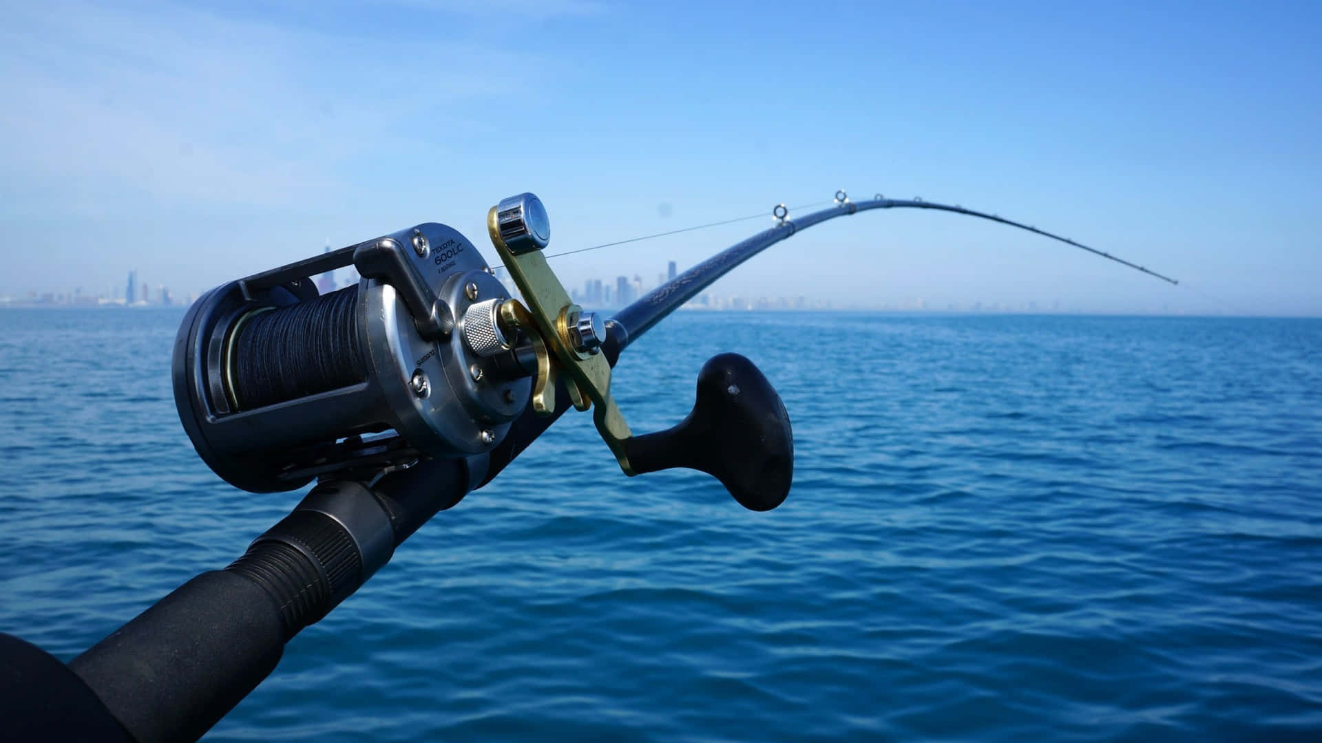 Fishing Rod 桌布 壁紙 — 彎曲的釣竿連捲線器在平靜的藍色水面上拋投，朝向晴朗天空下遠方的城市天際線。