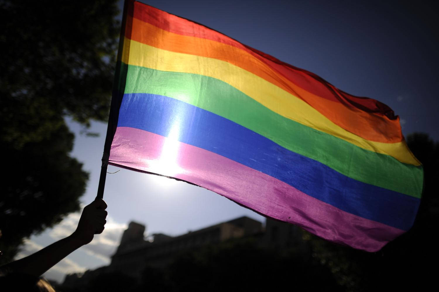 Pride Flag 桌布 壁紙 — 彩虹旗在明亮的陽光天空下自豪地飄揚，背景有樹木和建築物的剪影。