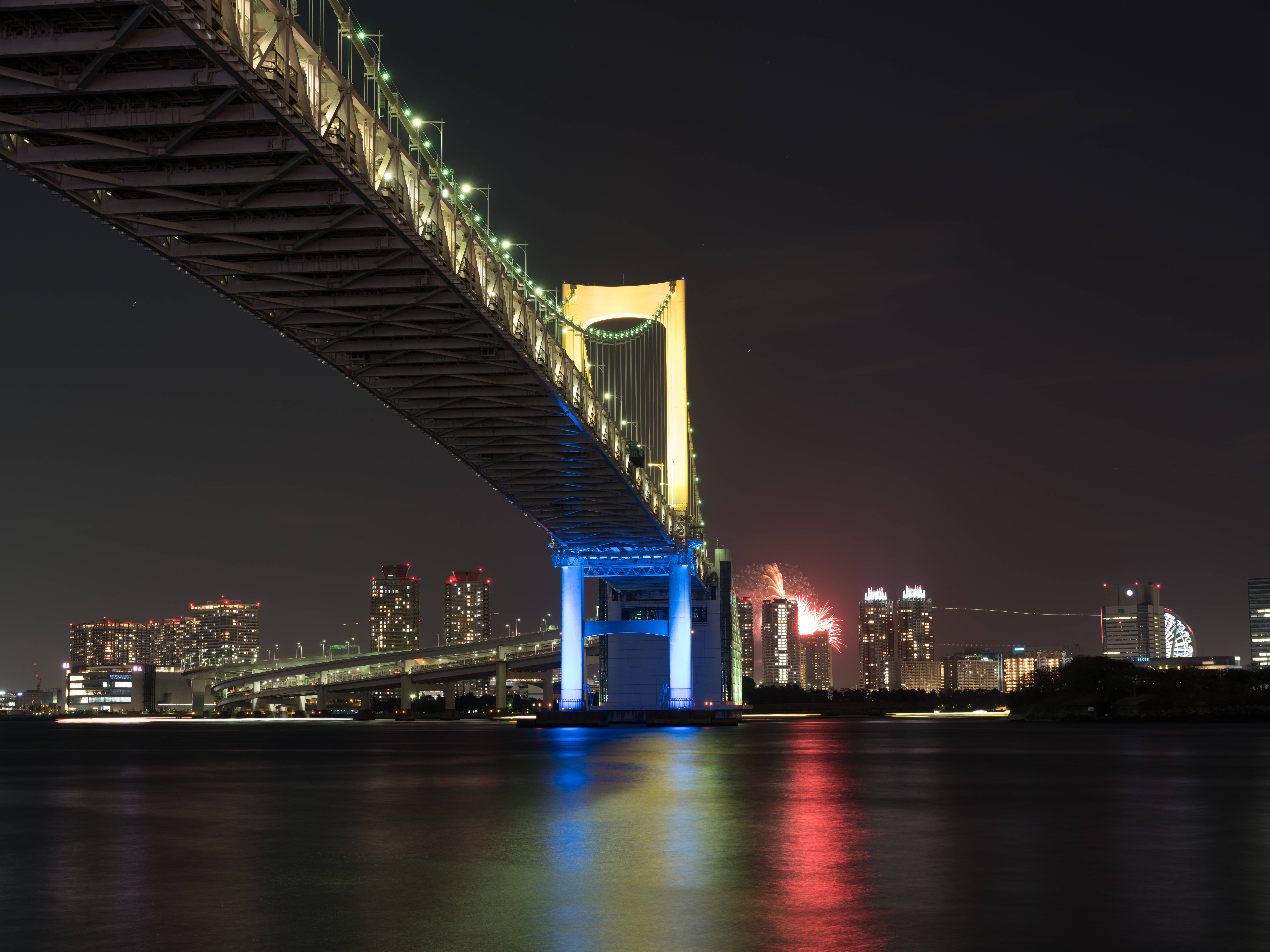 日本 桌布 壁紙 — 彩虹橋在夜間亮起，色彩繽紛的煙火在東京天際線上綻放，倒映在平靜的水面上。