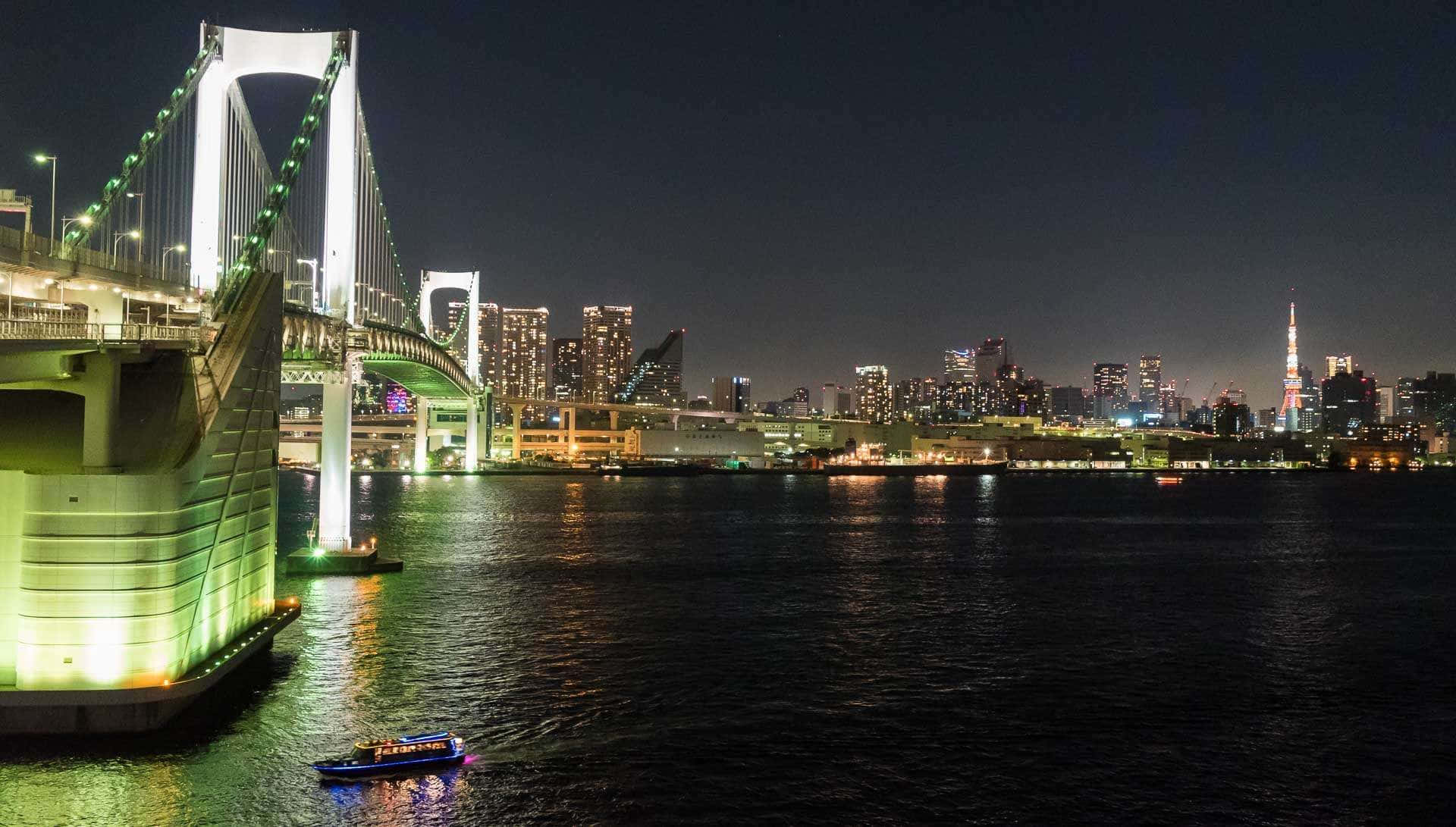 Rainbow Bridge 桌布 壁紙 — 彩虹橋在夜間照亮東京灣，城市燈光倒映在水面上，一艘小船駛過。