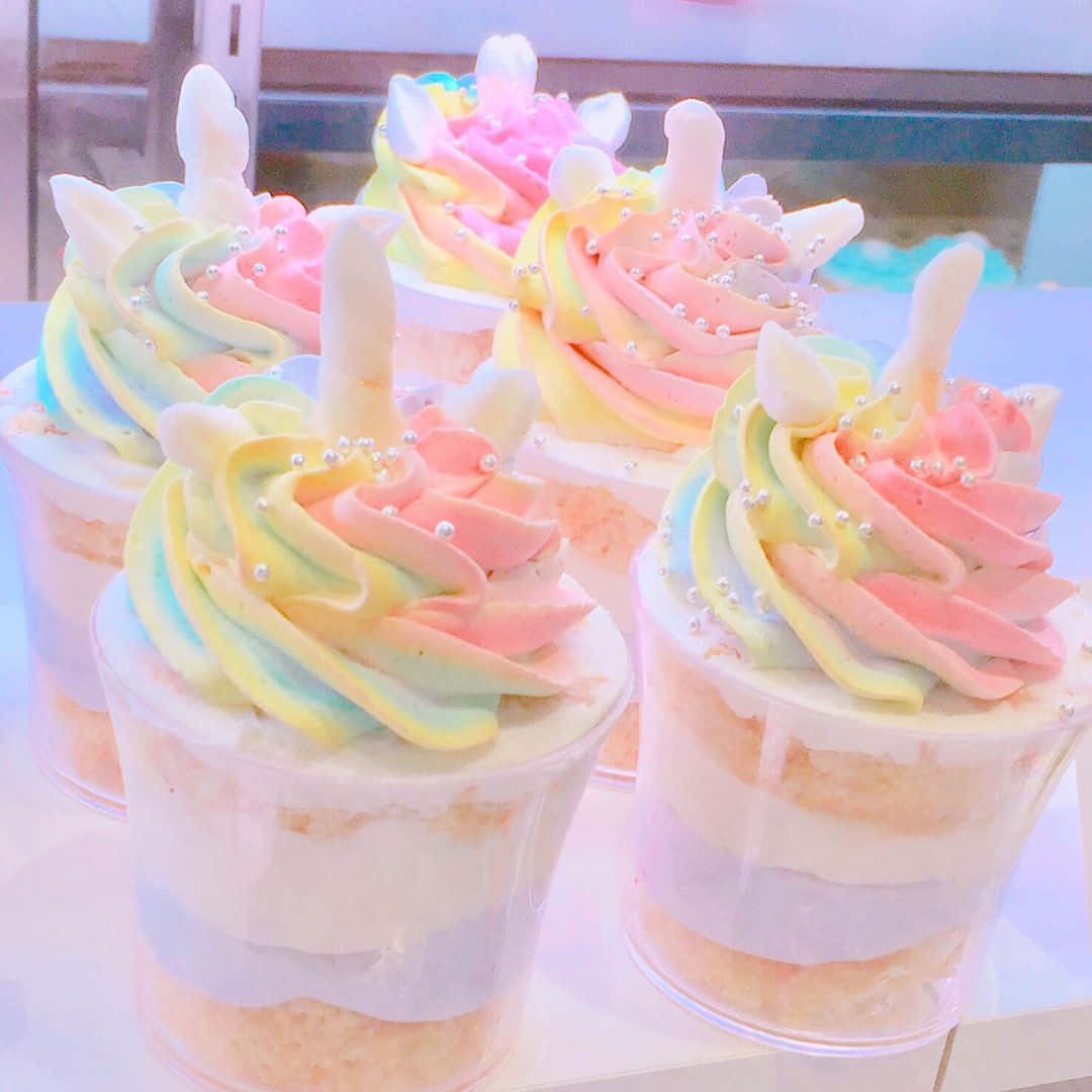 Kawaii Dessert 桌布 壁紙 — 彩虹漩渦獨角獸杯子蛋糕，搭配粉彩糖霜和珍珠糖粒，放在透明杯子裡，陳列在明亮的櫃檯上。