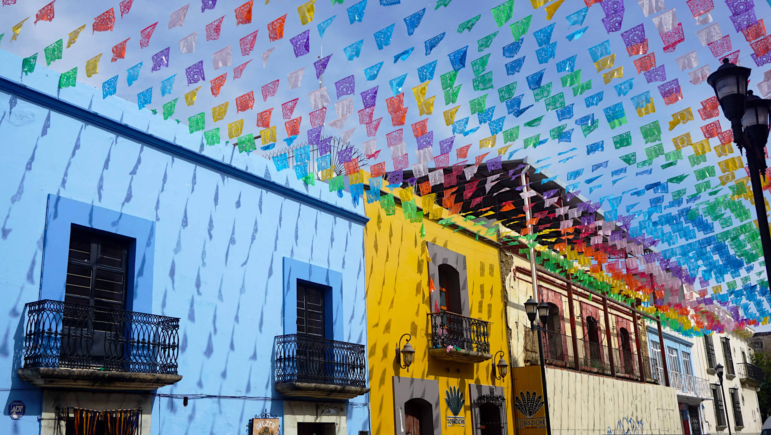 墨西哥 桌布 壁紙 — 彩虹色調的鮮豔 papel picado 橫幅掛在陽光明媚的街道上，街道兩旁是色彩繽紛的殖民時期建築。