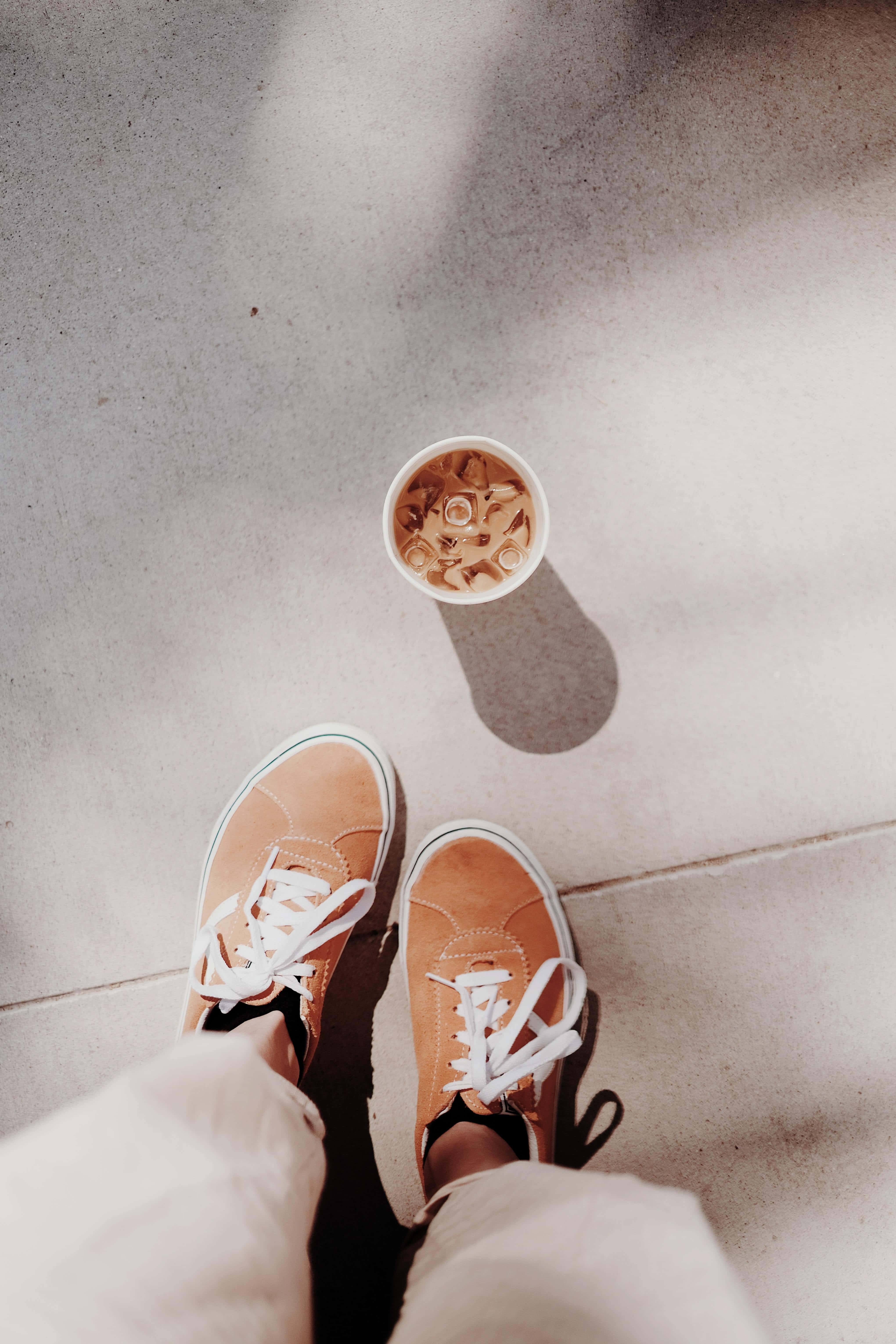 Iced Coffee 桌布 壁紙 — 從上方俯瞰，一雙淺褐色運動鞋和一杯冰咖啡放在陽光照射的混凝土地板上，投下柔和陰影。