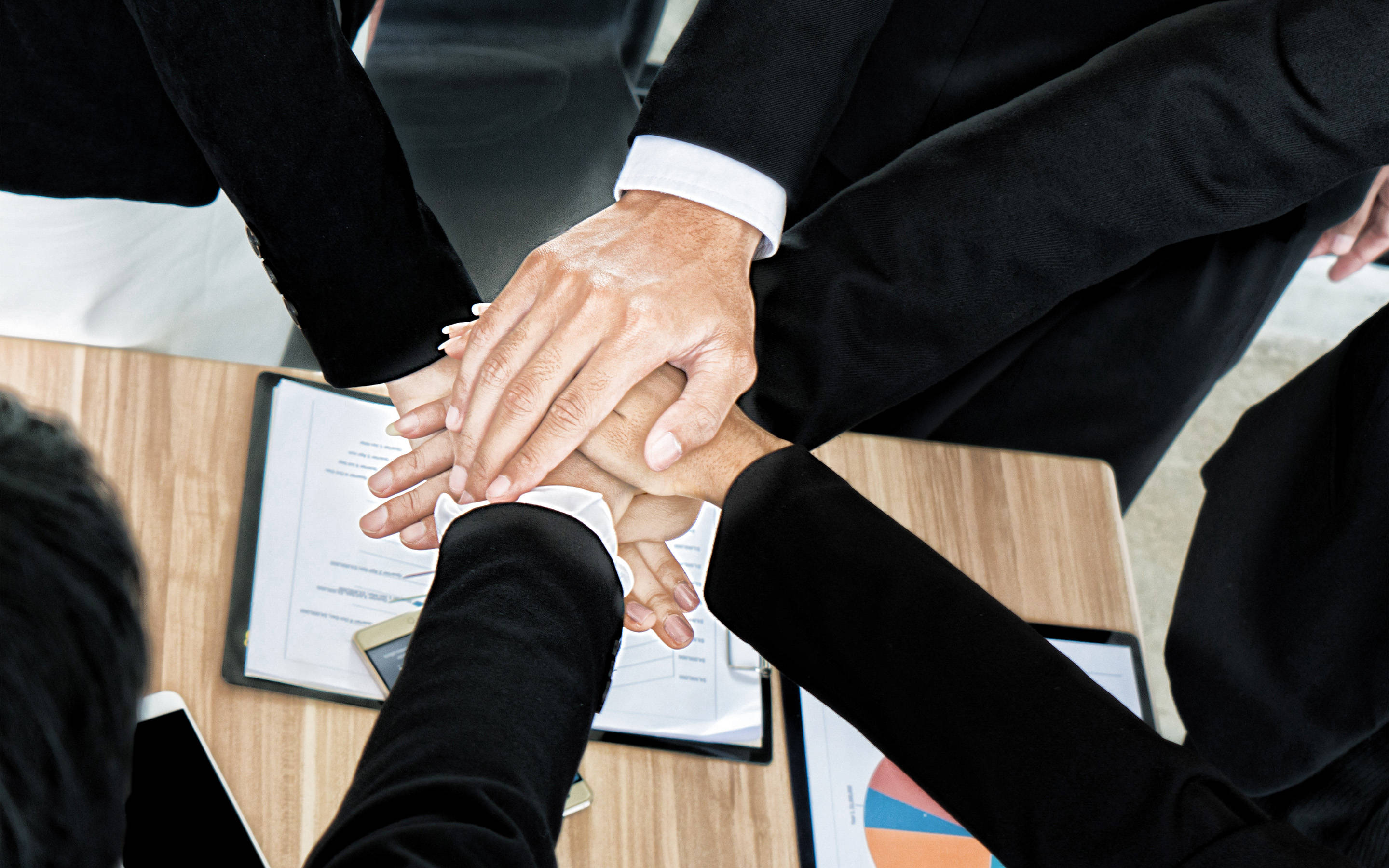 Teamwork 桌布 壁紙 — 從上方俯瞰，不同背景的商業人士在放有文件和圖表的木桌上團結疊手。
