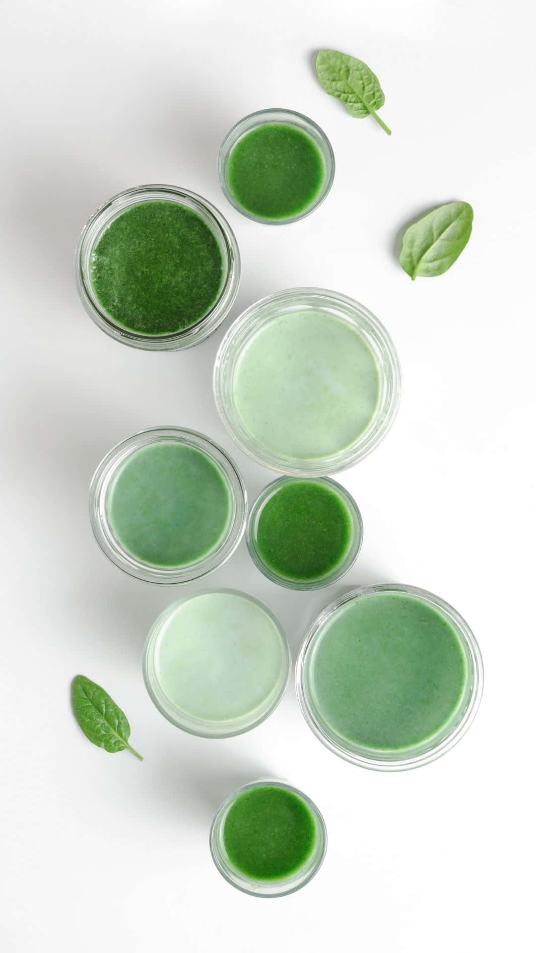 Sage 桌布 壁紙 — 從上方俯瞰，玻璃罐中裝有各種綠色冰沙，散落著新鮮菠菜葉，置於明亮的白色表面上。