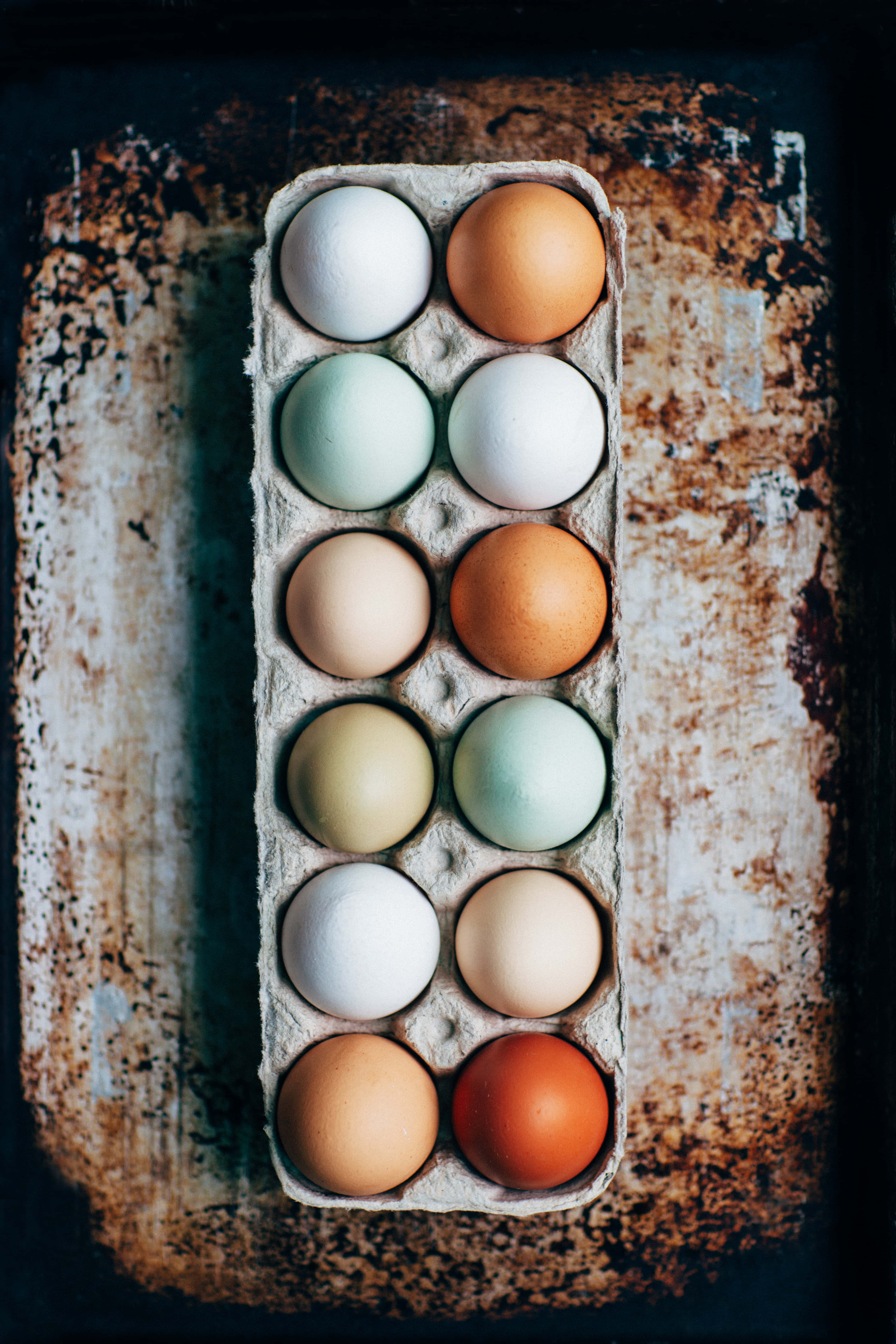 Eggs 桌布 壁紙 — 從上方俯瞰一個紙板雞蛋盒，裡面裝著十二顆色彩繽紛的雞蛋，放在一個生鏽且風化的表面上。