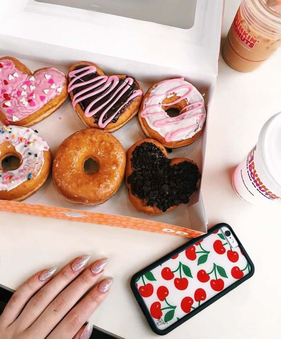 Dunkin Donuts 桌布 壁紙 — 從上方俯瞰一個裝滿心形和糖霜甜甜圈的Dunkin' Donuts盒子，旁邊有櫻桃圖案的手機殼和冰咖啡。