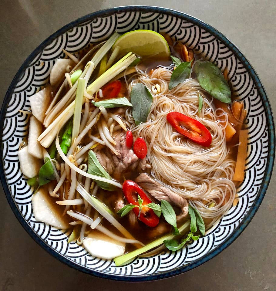 越南 桌布 壁紙 — 從上方俯瞰一碗冒煙的湯麵，碗內有米粉、豆芽、羅勒、萊姆和紅辣椒片，盛載於有圖案的陶瓷碗中。