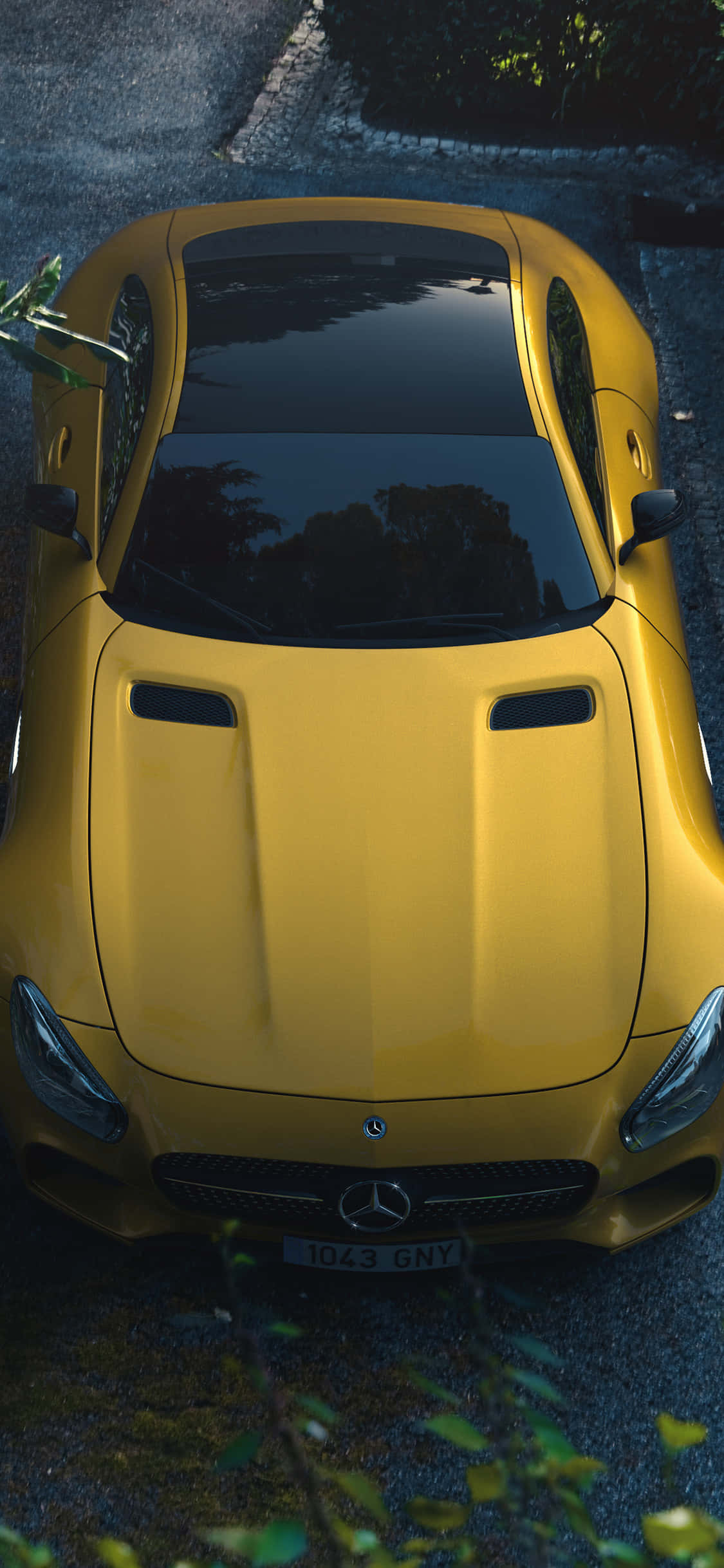 iPhone X Amg Gt-r 桌布 壁紙 — 從上方俯瞰一輛充滿活力的黃色 Mercedes-AMG GT 跑車停泊在鵝卵石街道上，周圍有綠色植物。