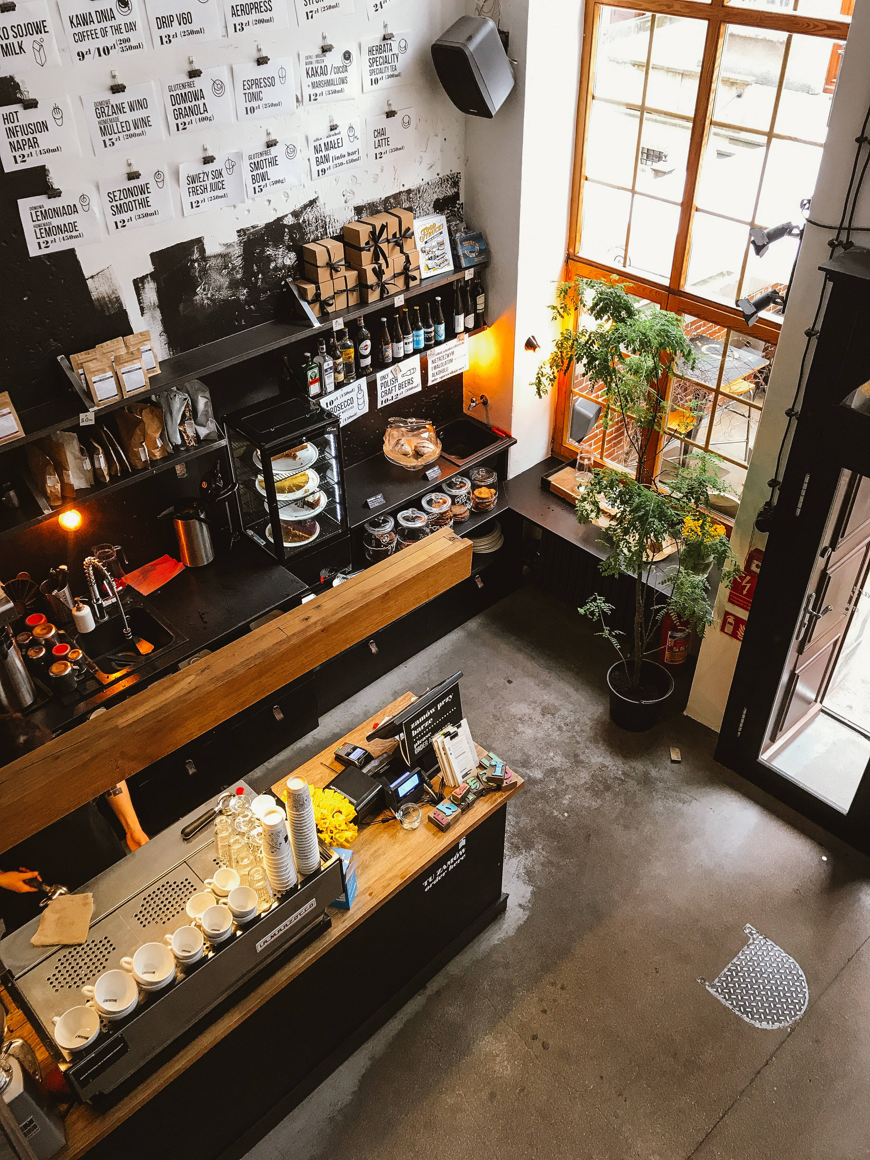 Coffee Shop 桌布 壁紙 — 從上方俯瞰一間現代咖啡店，設有木製櫃檯、黑色層架，以及靠近大窗戶的盆栽植物。