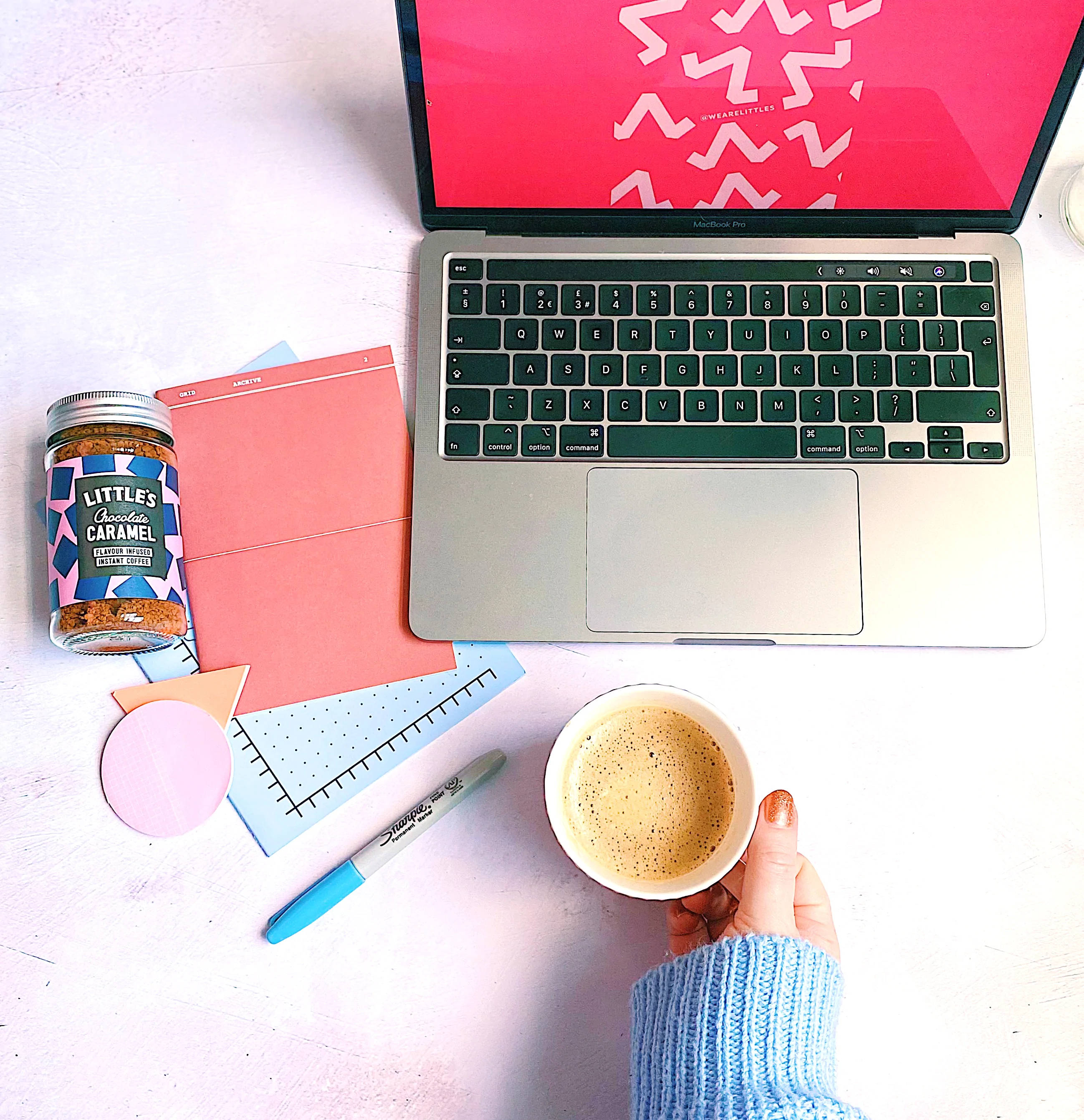 Coffee Aesthetic 桌布 壁紙 — 從上方俯瞰一隻手在白色桌子上拿著咖啡，旁邊有筆記型電腦、焦糖罐和彩色文具。