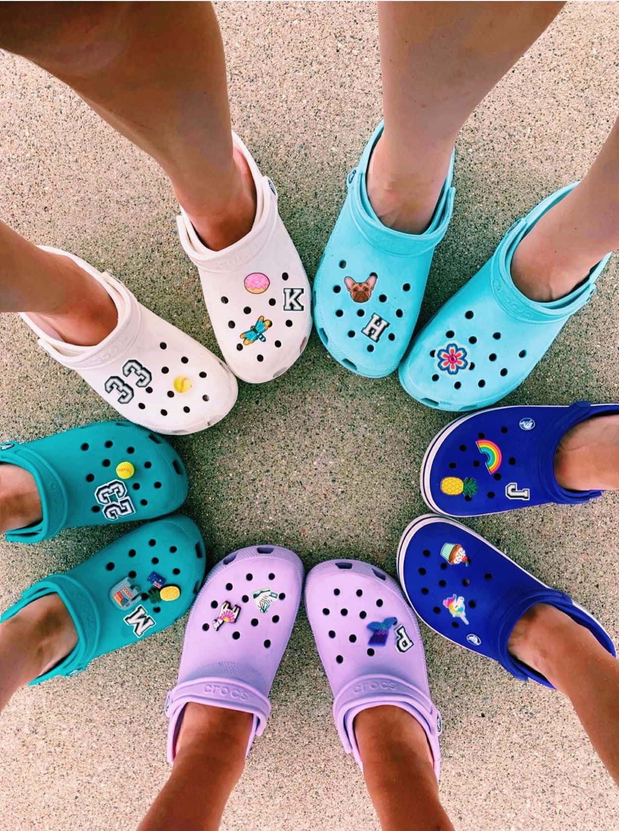 Crocs 桌布 壁紙 — 從上方俯瞰六個人的腳，穿著色彩繽紛的Crocs，在斑點路面上排成一個圓圈，每隻鞋都裝飾著獨特的飾物。