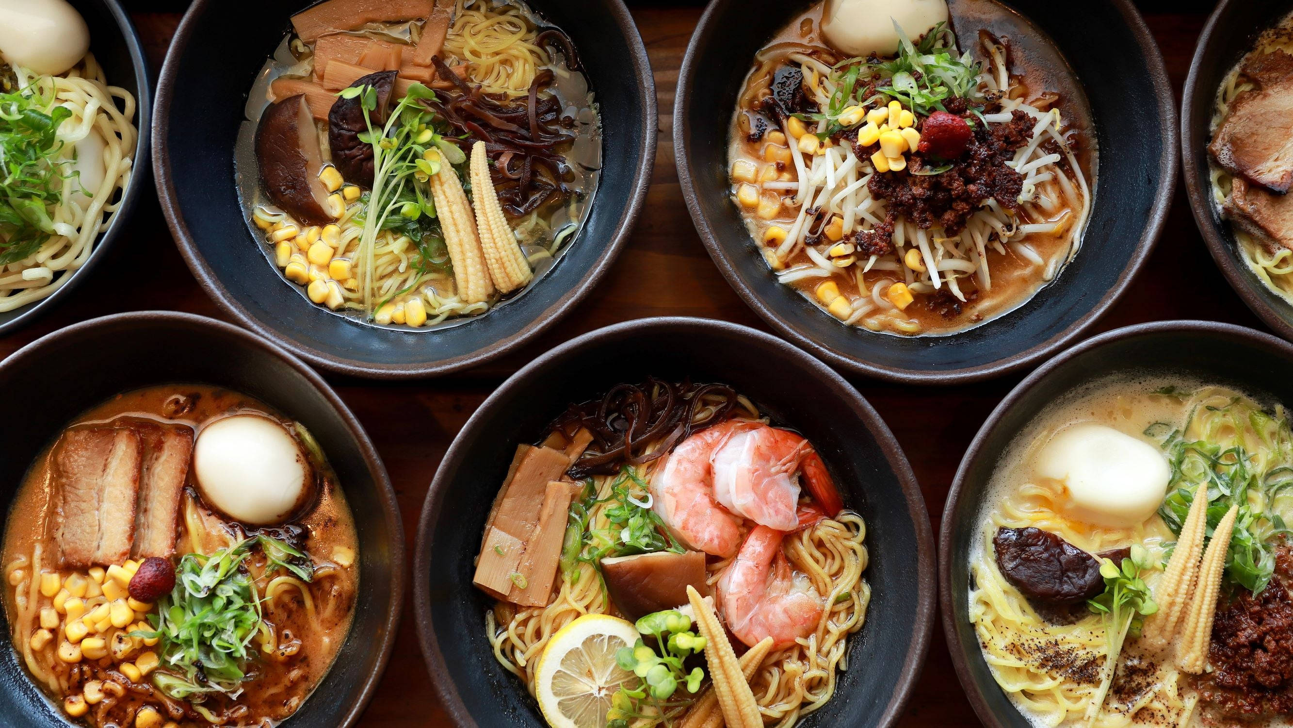 Ramen 桌布 壁紙 — 從上方俯瞰六碗拉麵，配料多樣，包括蝦、玉米和半熟蛋，置於木桌上。