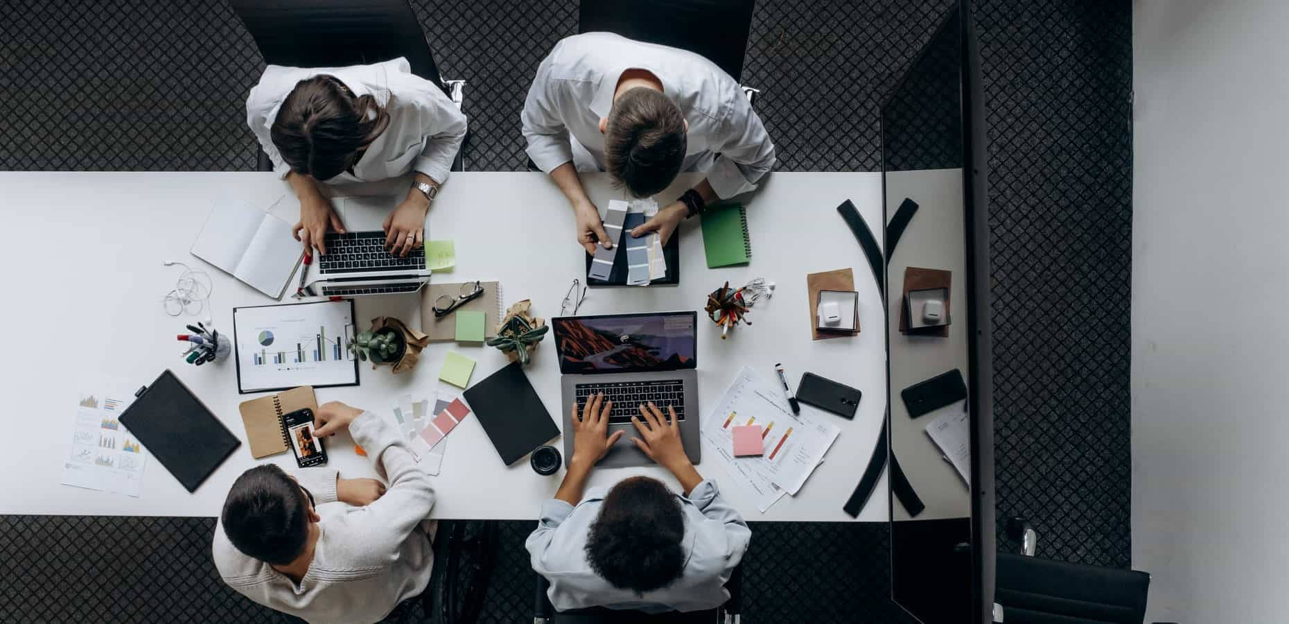Working 桌布 壁紙 — 從上方俯瞰四人在現代辦公室的白桌上協作，桌上放有手提電腦、筆記本和設計材料。