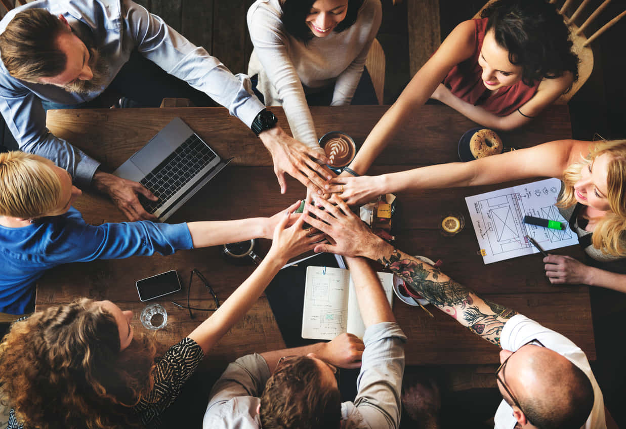 Teamwork 桌布 壁紙 — 從上方俯瞰多元團隊圍繞木桌，桌上放有筆記型電腦和咖啡，團隊團結一致疊手。