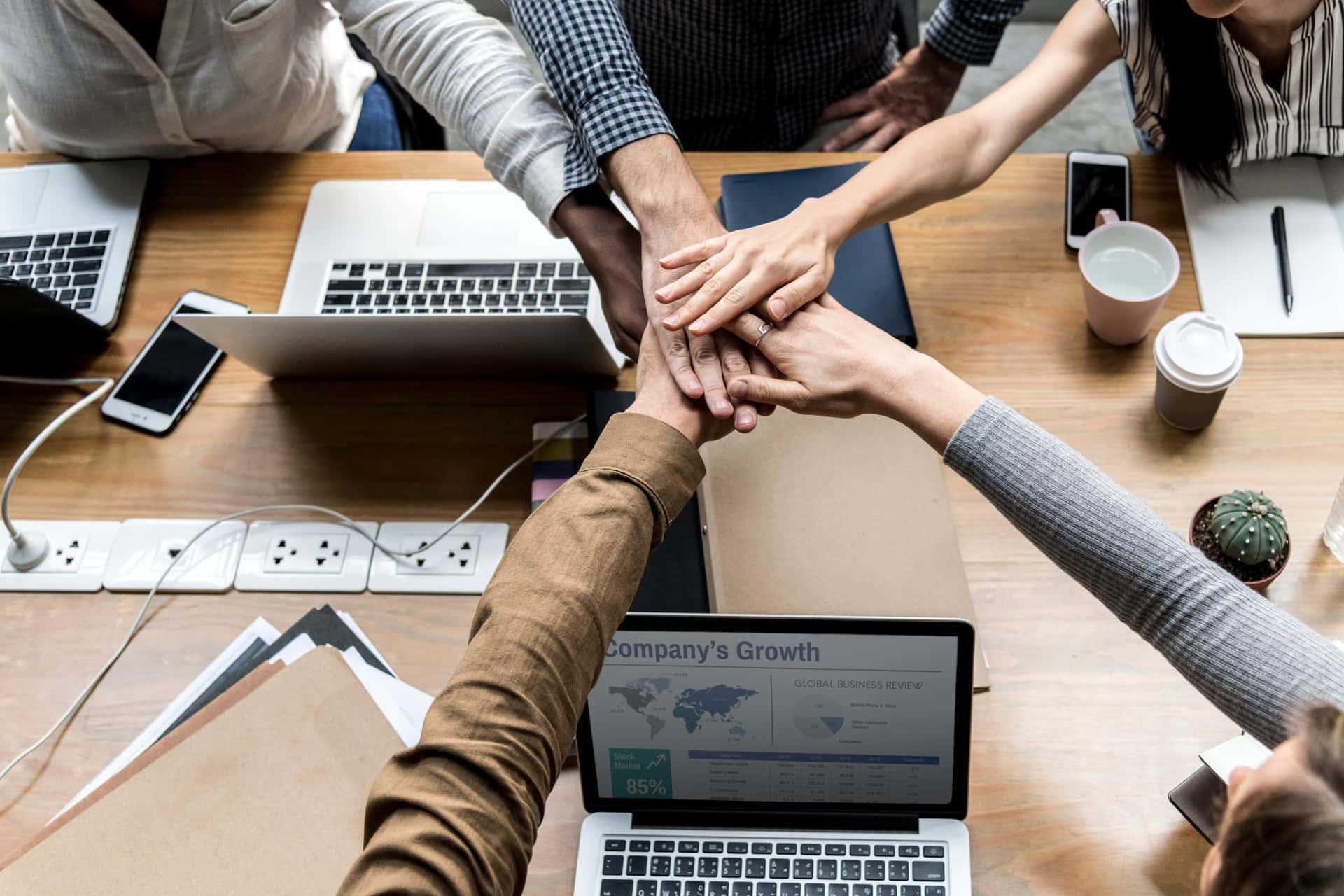 Teamwork 桌布 壁紙 — 從上方俯瞰多元團隊圍繞木桌團結疊手，桌上放有筆記型電腦和商業圖表。