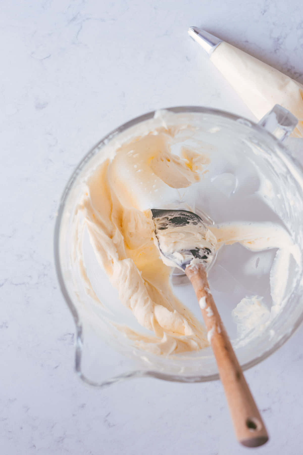 Buttercream 桌布 壁紙 — 從上方俯瞰大理石表面上玻璃攪拌碗中的奶油淡黃色糖霜，配有木製刮刀和裱花袋。