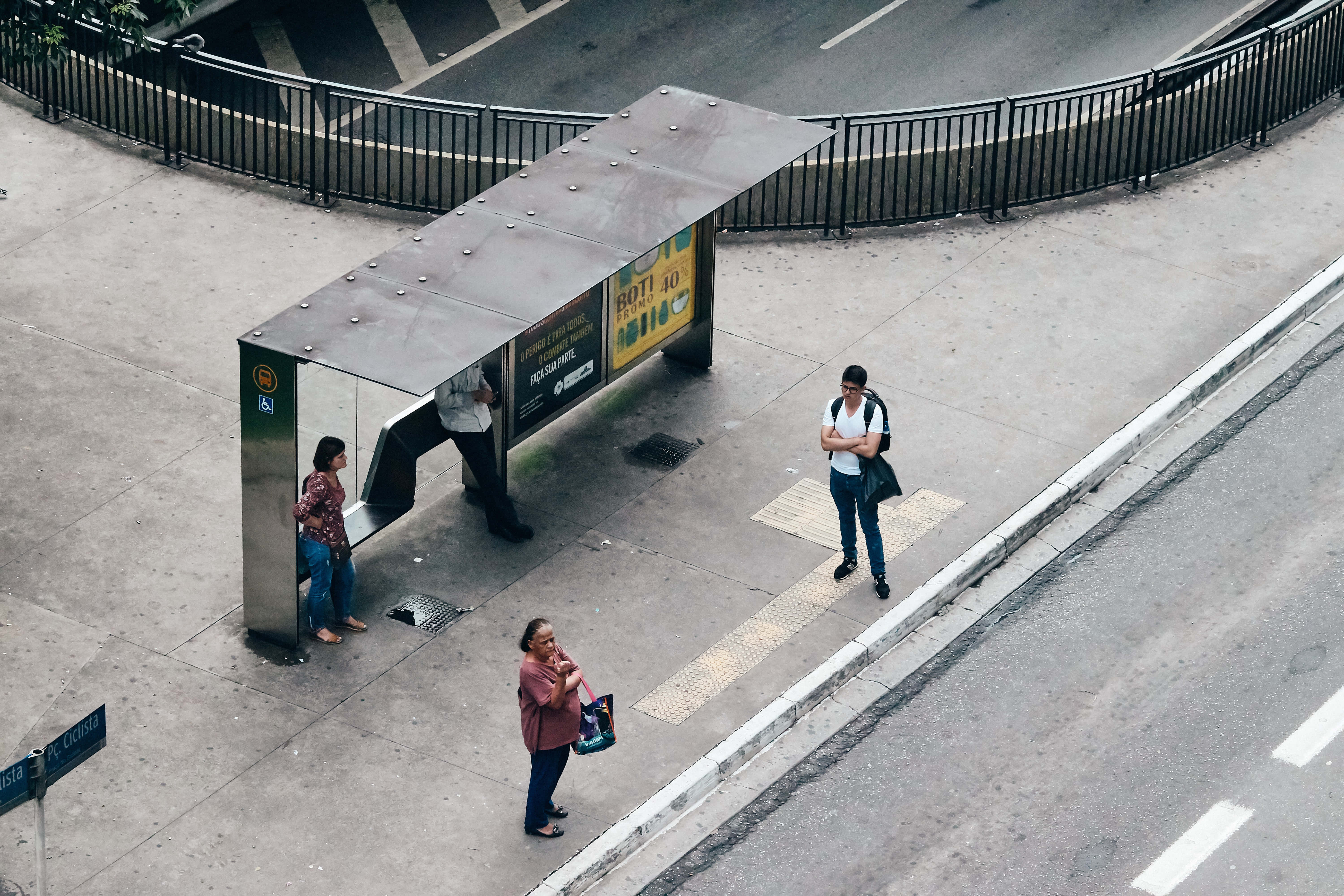 Waiting 桌布 壁紙 — 從上方俯瞰市區公車站，三人正在人行道上等候，旁邊是帶有白色車道標線的瀝青道路。