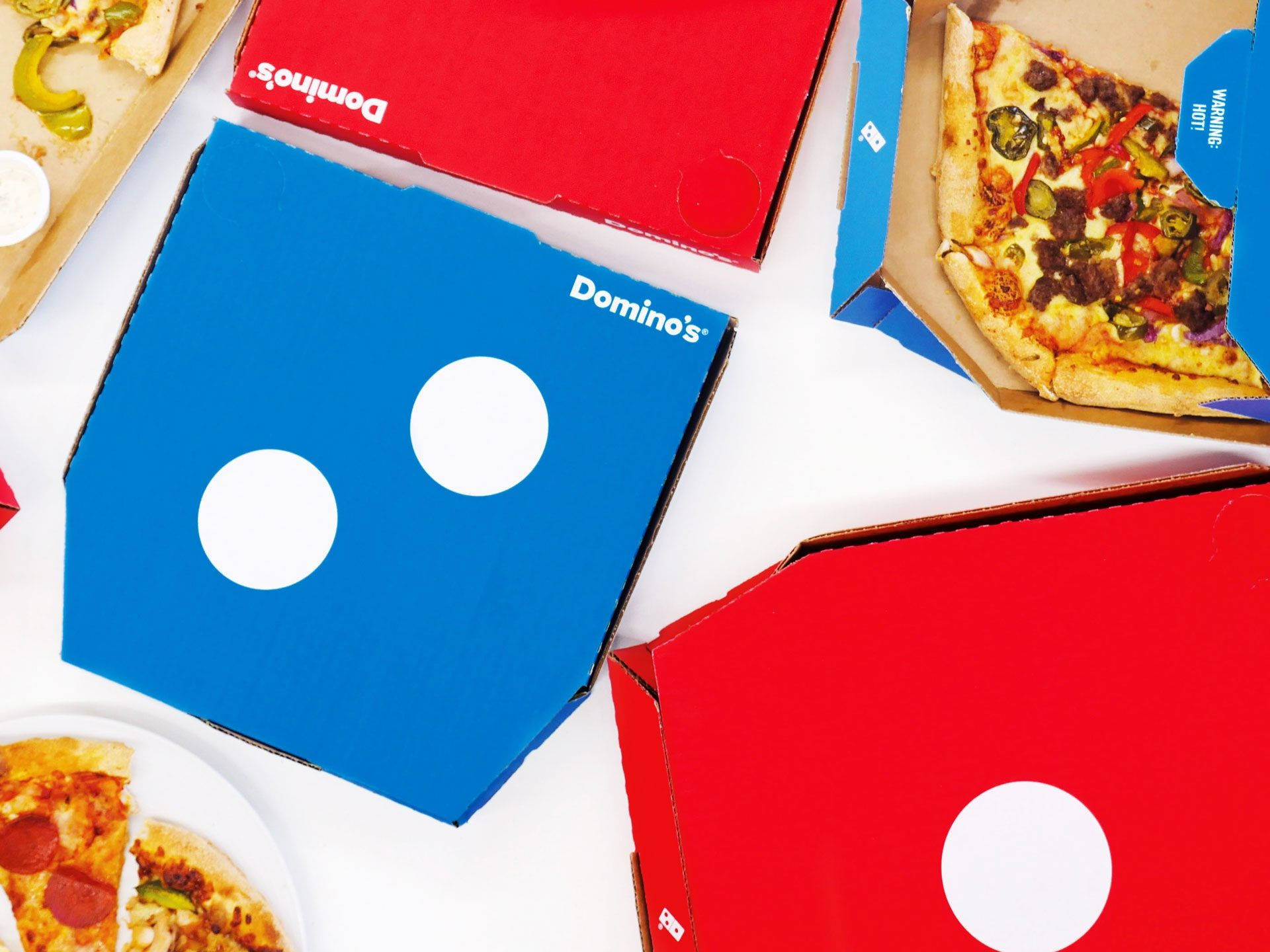 Dominos Pizza 桌布 壁紙 — 從上方俯瞰打開的Domino's披薩盒，上面有色彩繽紛的配料和品牌標誌，放在潔淨的白色表面上。