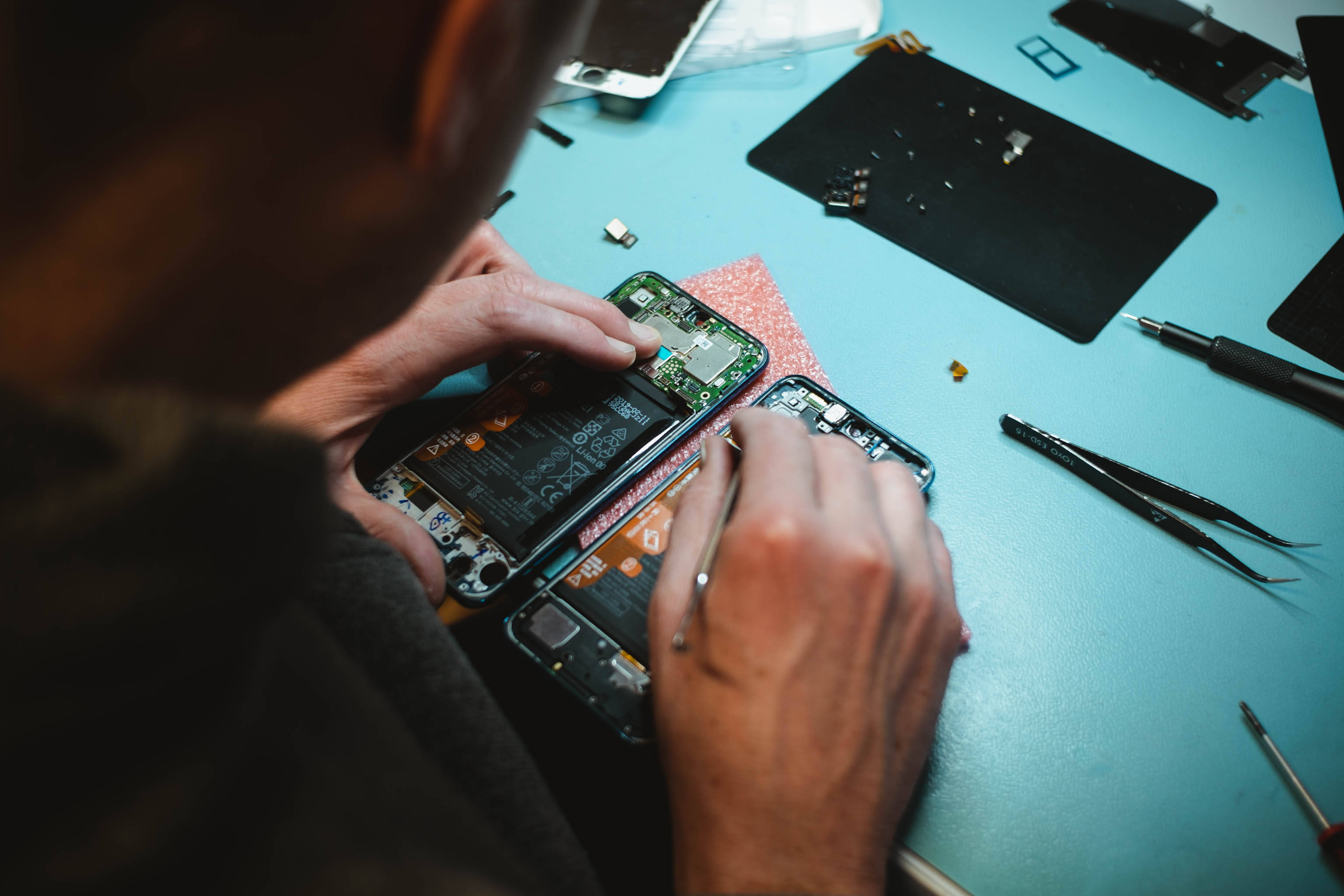手機破裂 桌布 壁紙 — 從上方俯瞰技術人員在藍色工作台上用工具仔細維修智慧型手機電路板。