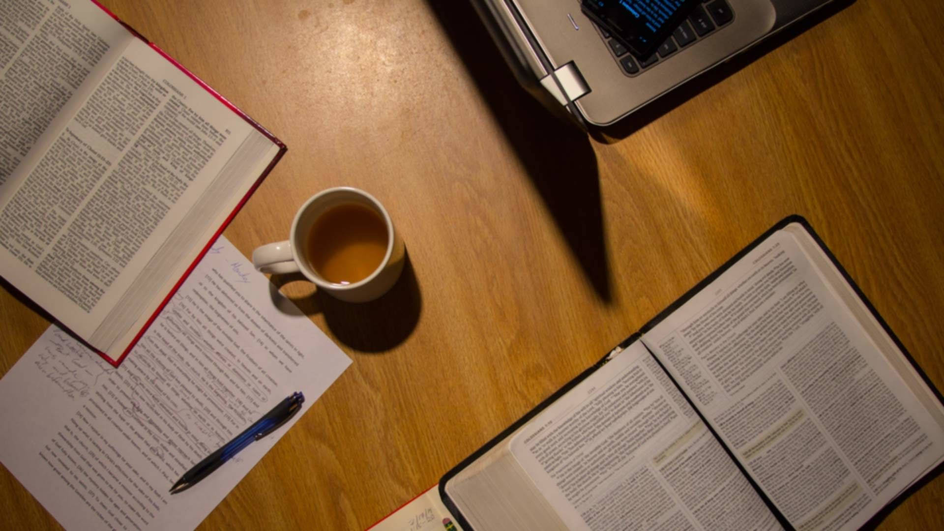 Studying 桌布 壁紙 — 從上方俯瞰木製書桌，上面有打開的書本、咖啡杯和筆記型電腦，沐浴在溫暖的環境光中。