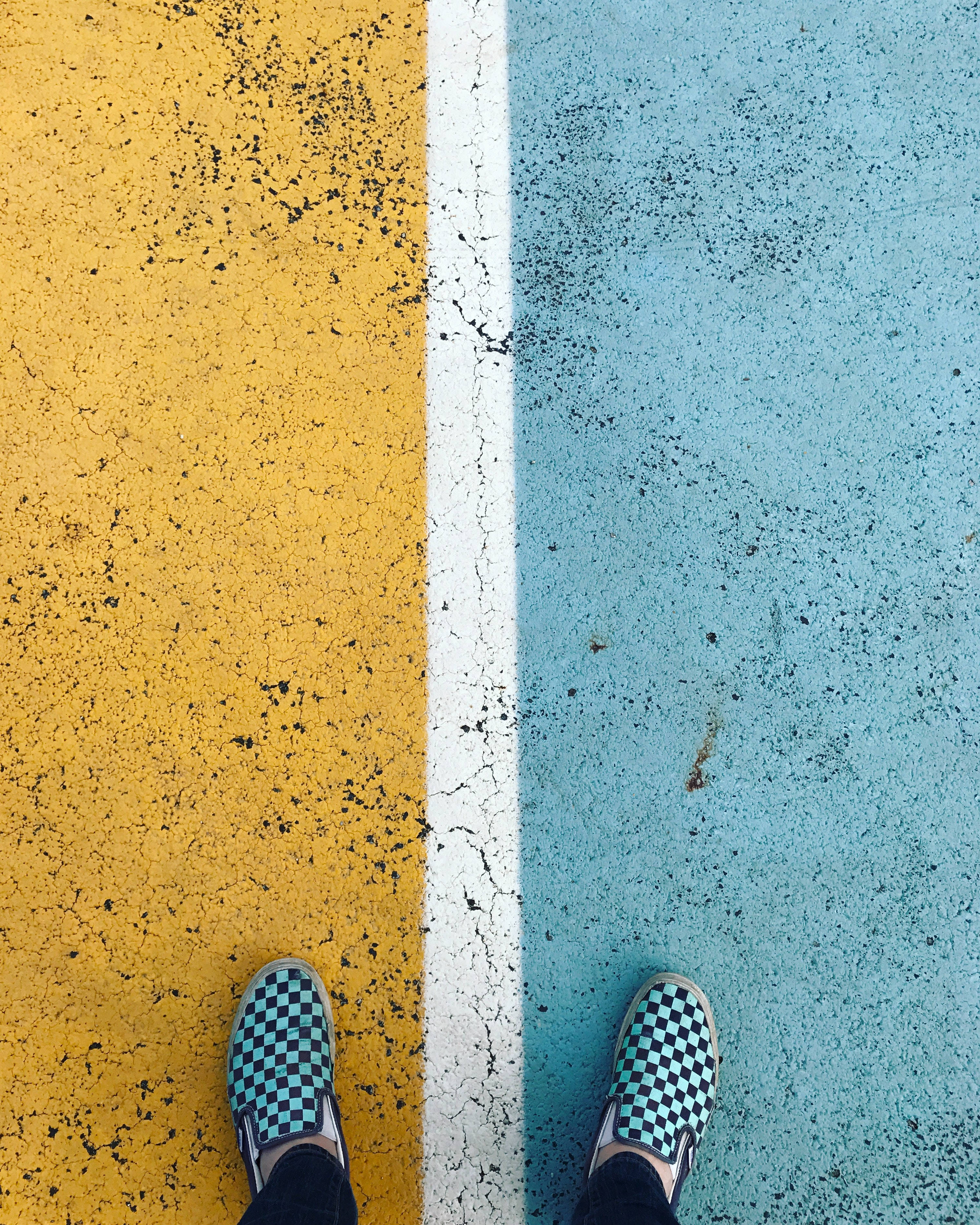 混凝土 桌布 壁紙 — 從上方俯瞰格紋 slip-on 鞋站在一條裂開的白線上，該線分隔黃色和藍色的瀝青路面。
