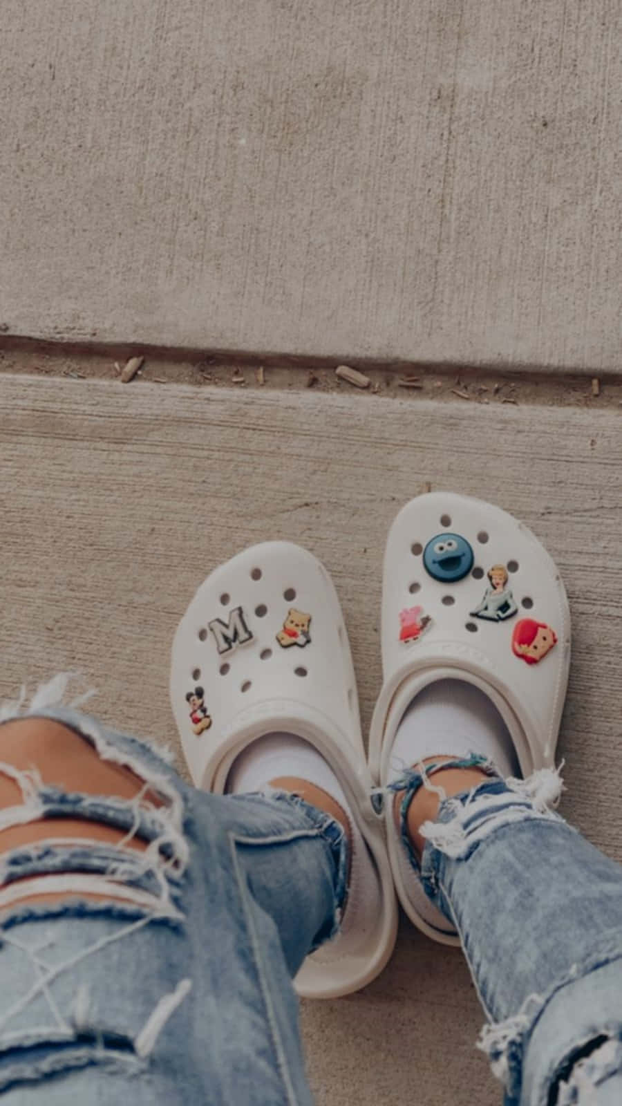 Crocs 桌布 壁紙 — 從上方俯瞰白色Crocs鞋，飾有色彩繽紛的角色吊飾，搭配破洞牛仔褲，置於混凝土人行道上。